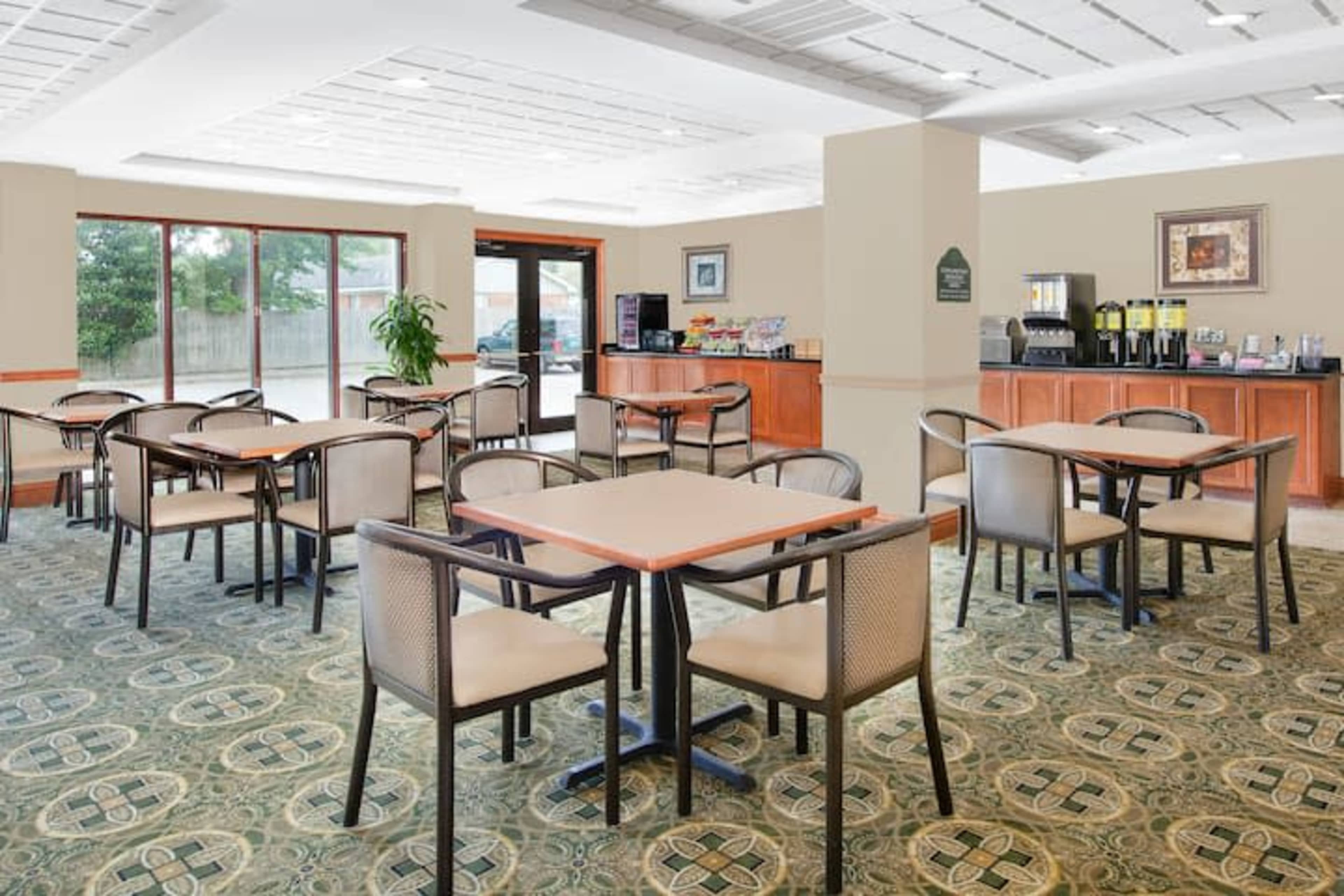 A brightly lit breakfast area with several tables and chairs arranged around a patterned carpet, alongside a counter featuring food and beverage offerings.