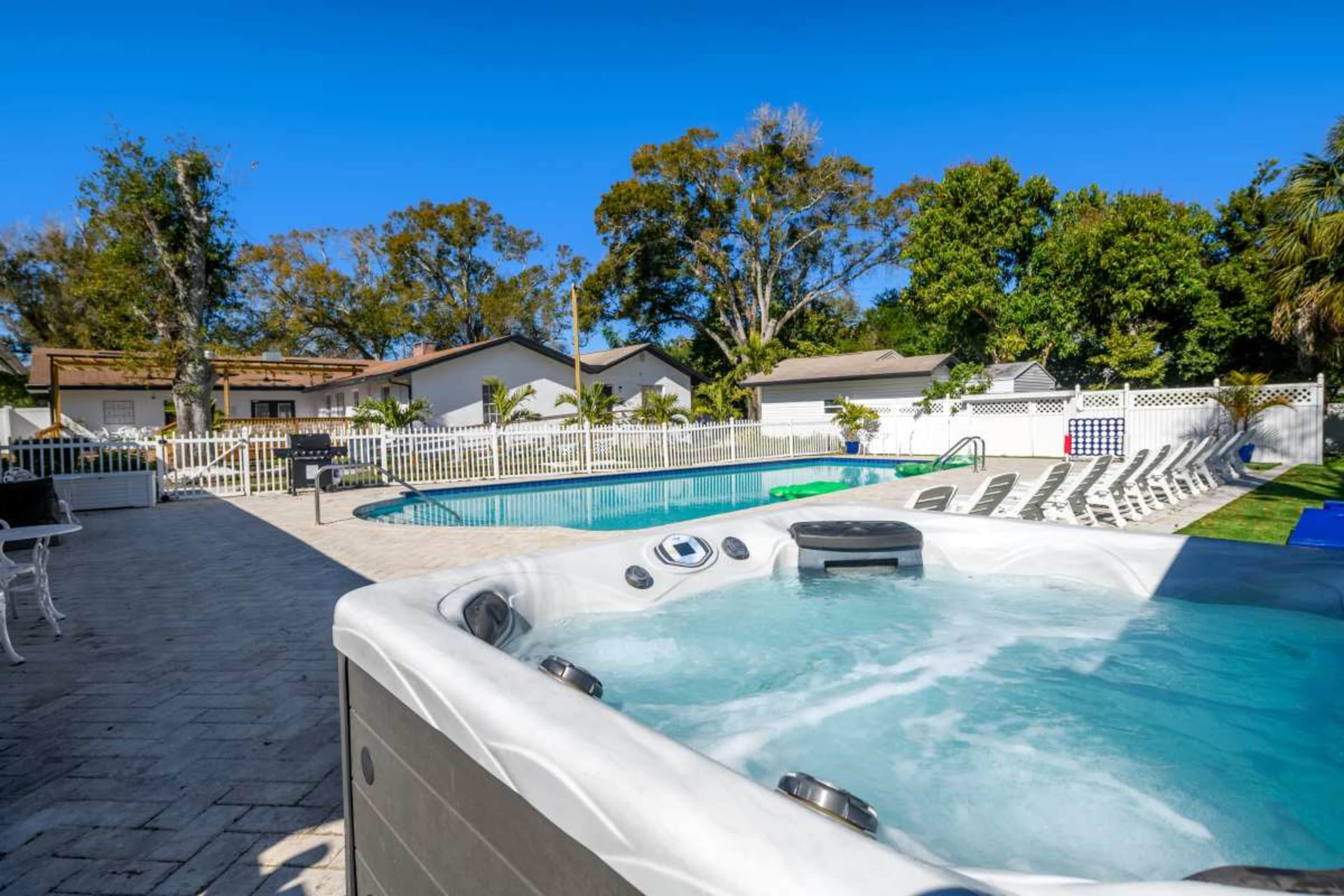 A hot tub is positioned in the foreground next to a swimming pool surrounded by lounge chairs and a white fence.