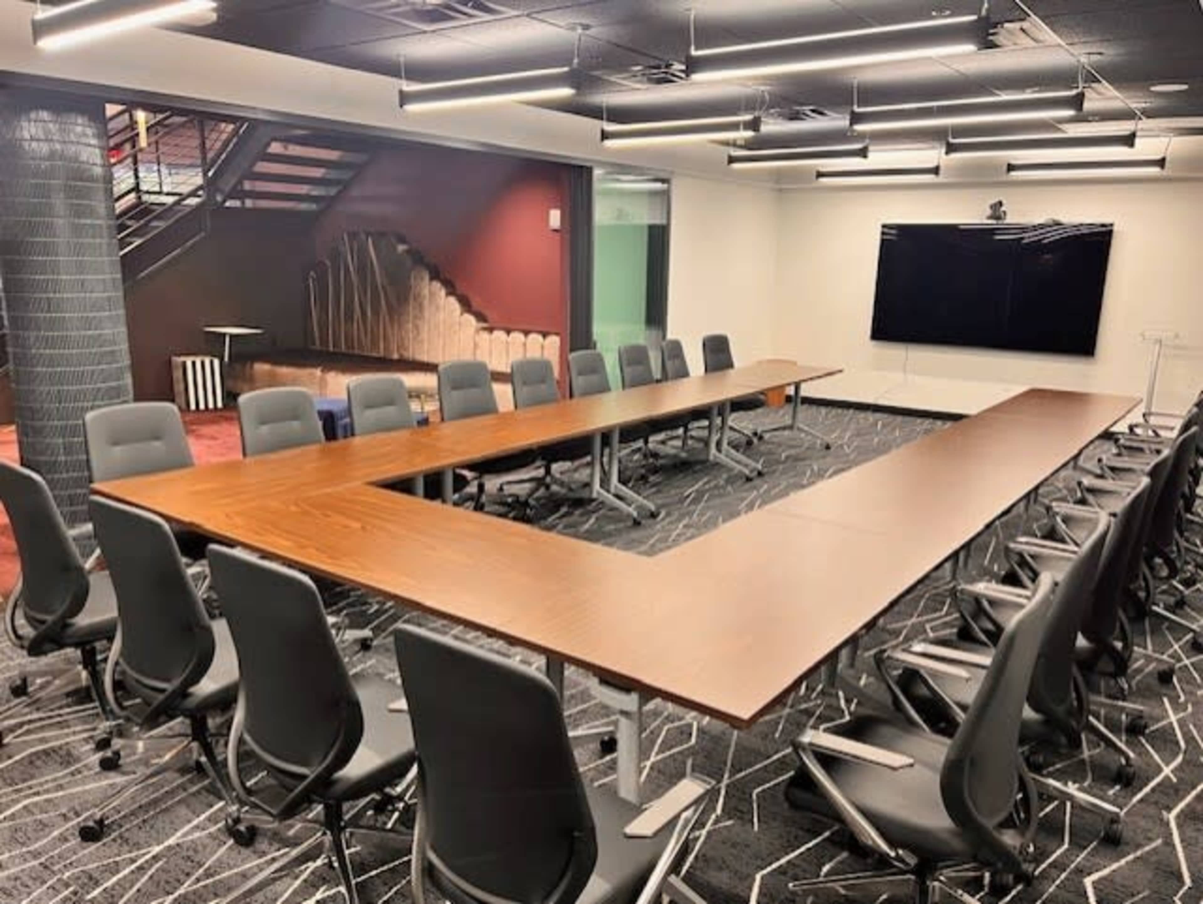 The image shows a modern conference room featuring a large, U-shaped table surrounded by ergonomic chairs, with a staircase and a large screen in the background.
