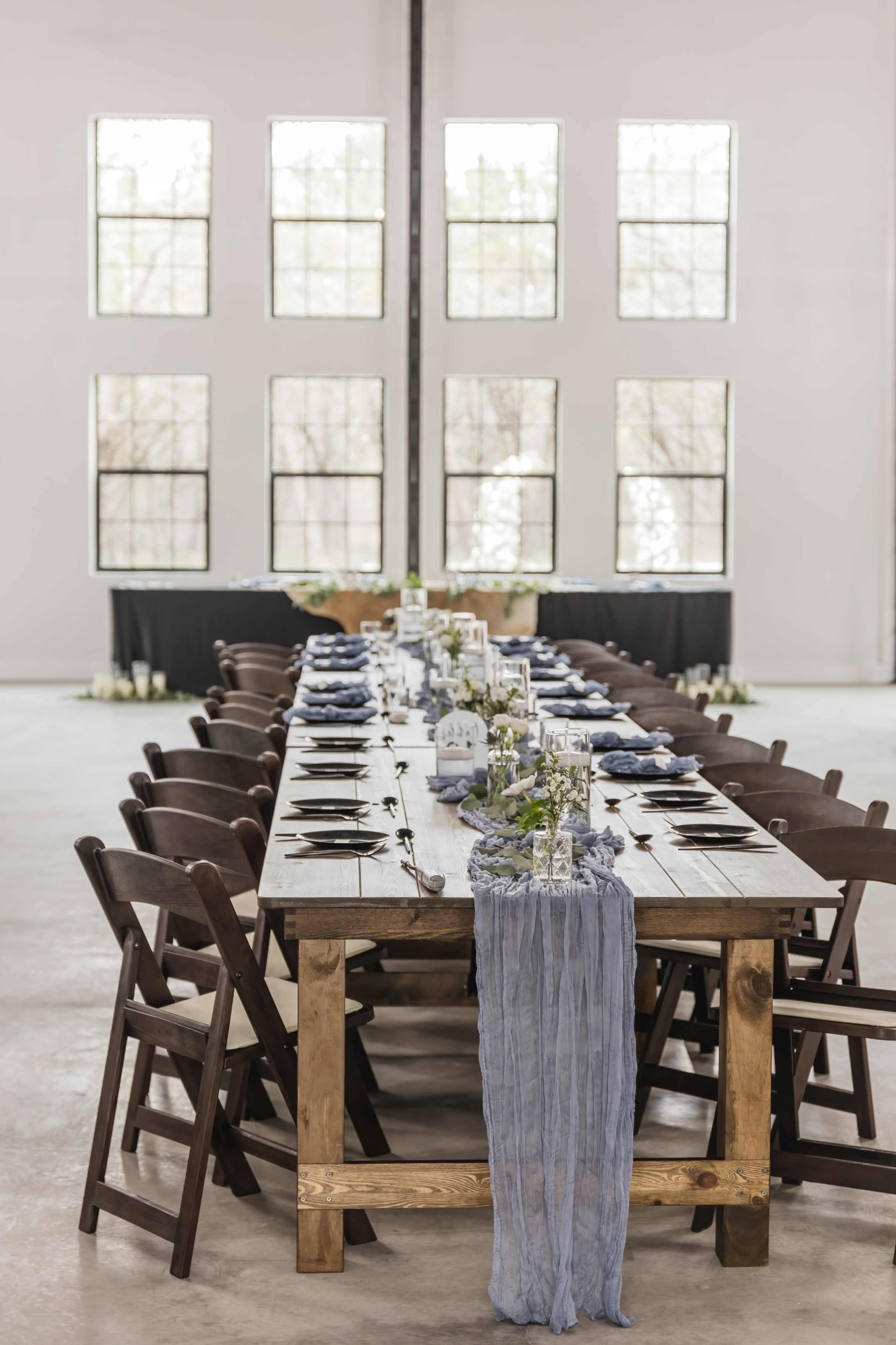 A long, rustic wooden dining table is set for a gathering, with elegant place settings and a blue runner, surrounded by dark chairs in a bright room featuring large windows.