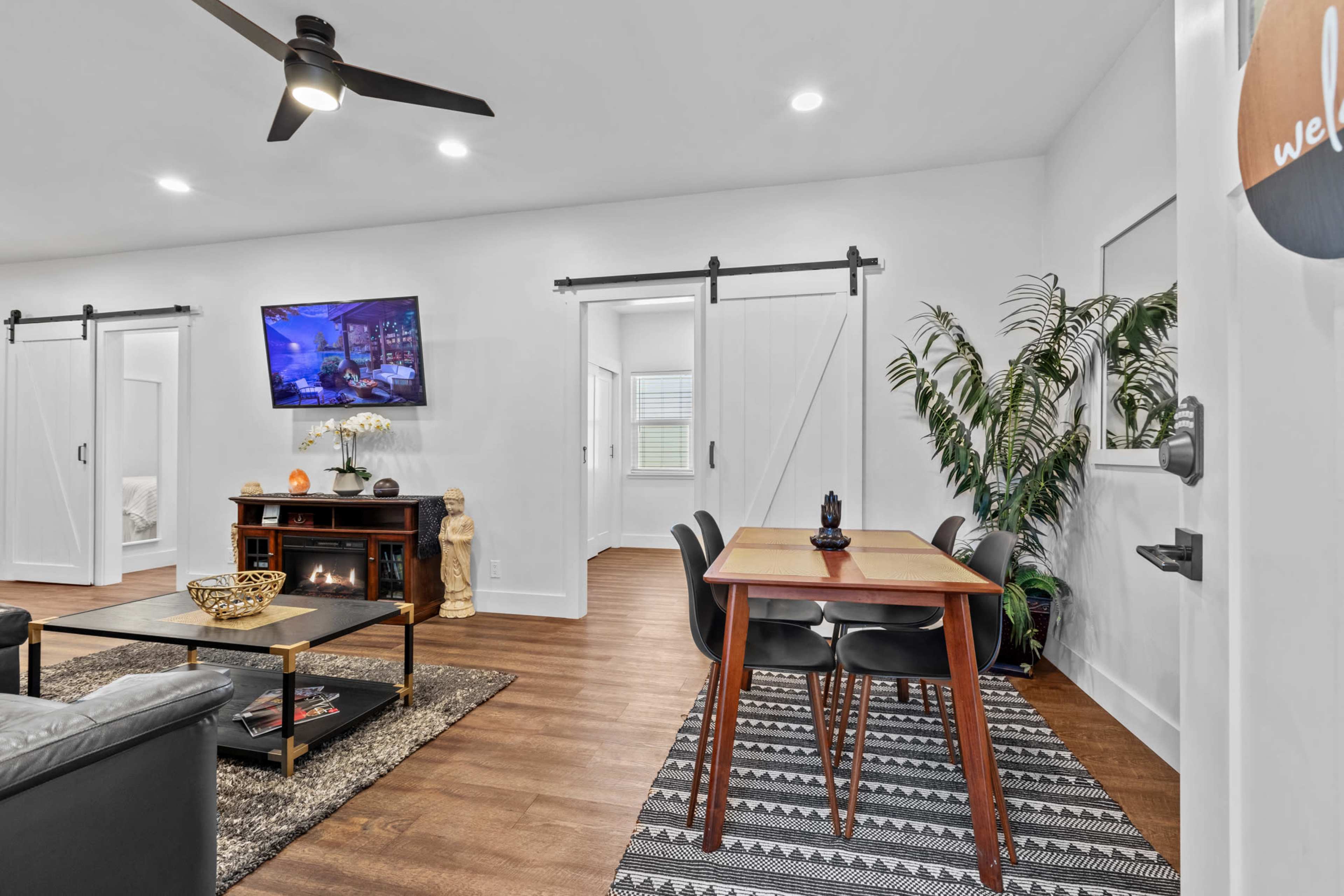 A modern living space features a dining area with a table and chairs, a black sofa, a flat-screen TV on the wall, and sliding barn doors leading to another room.
