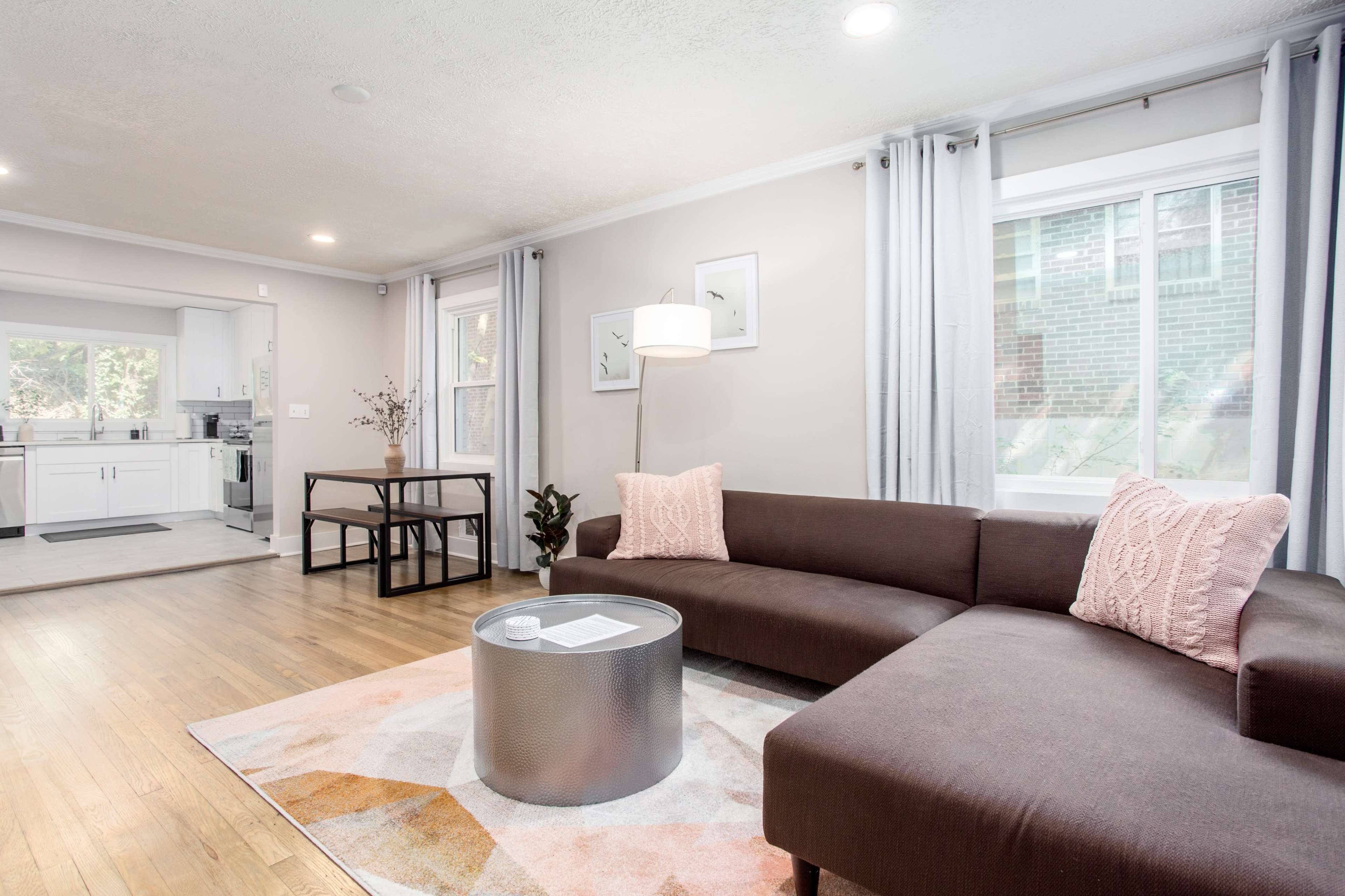 A modern living room features a brown sectional sofa, a round silver coffee table, and a small dining area, with large windows allowing natural light to fill the space.