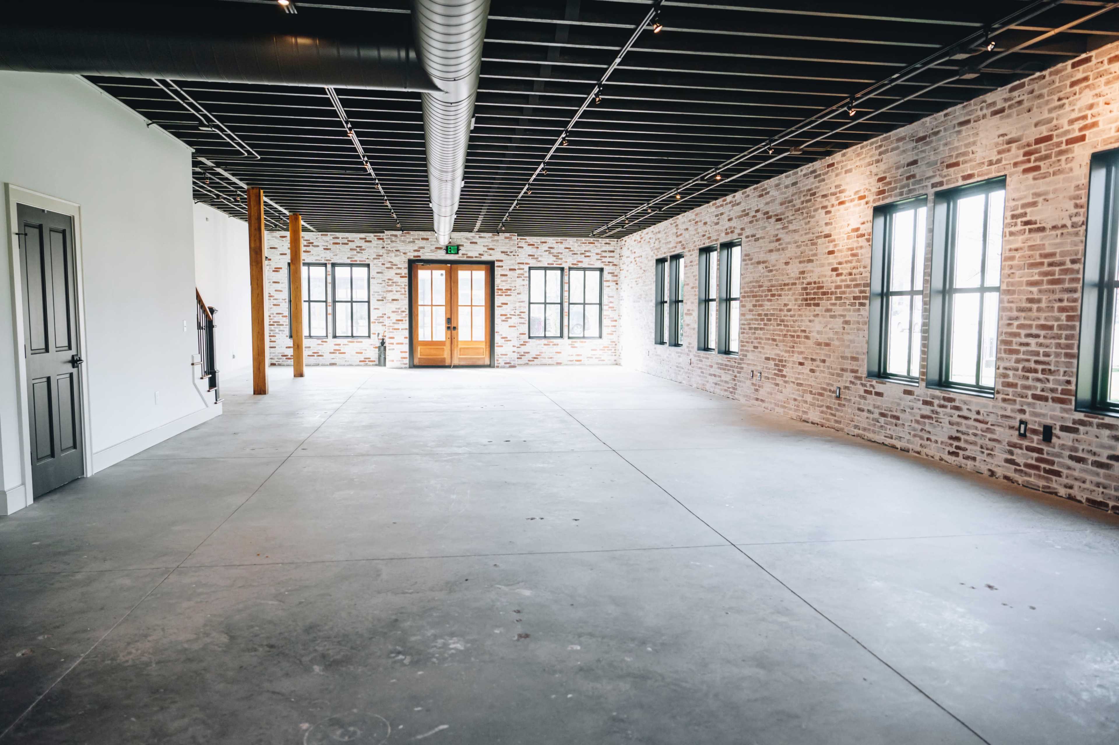 An empty interior space with exposed brick walls, large windows, and a concrete floor.