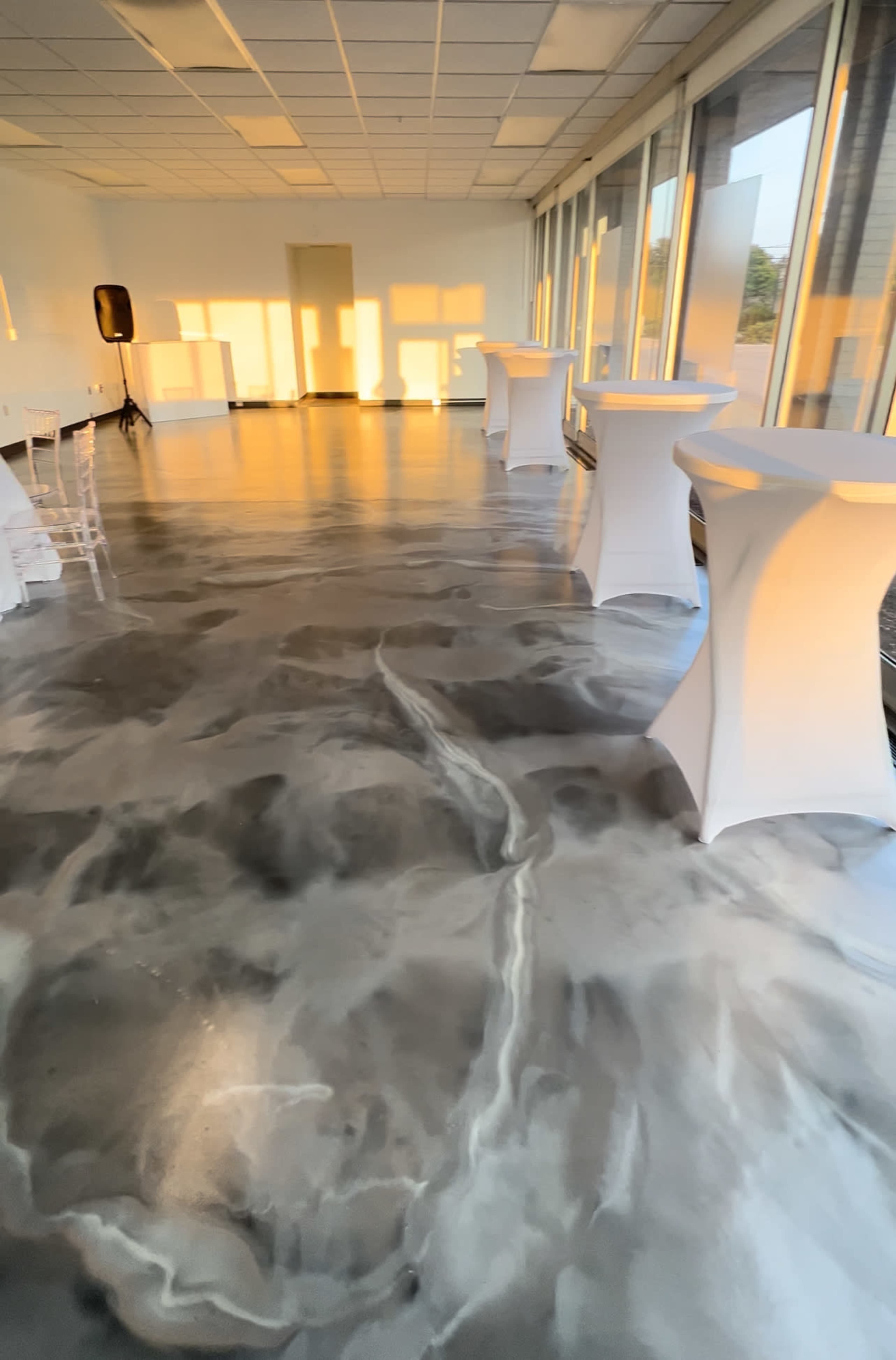 A modern event space with glossy, marbled flooring and several tall tables arranged along the windows, lit by warm afternoon sunlight.