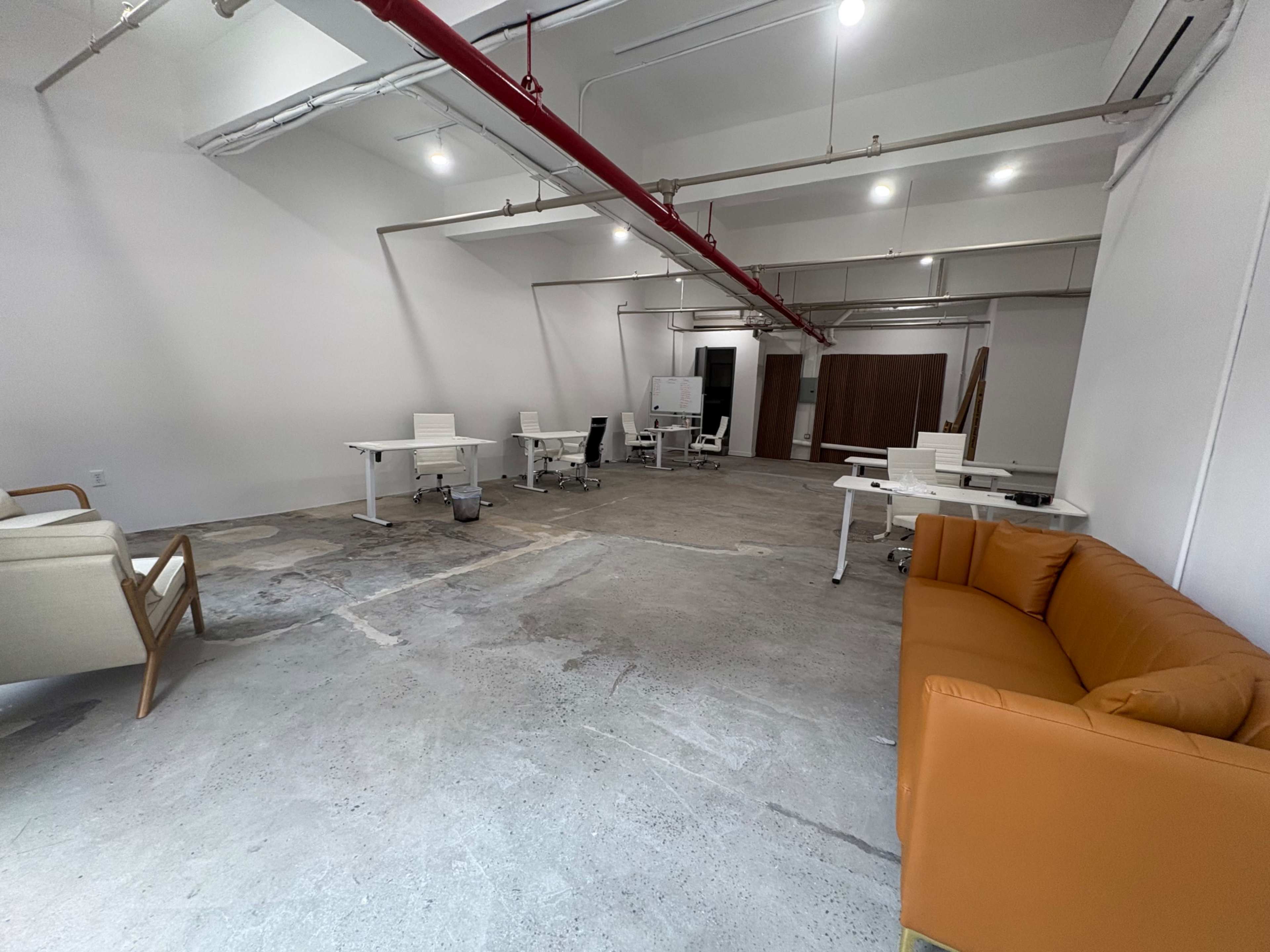 The image shows a spacious, minimally furnished office area with several desks and chairs, along with an orange sofa in the corner.