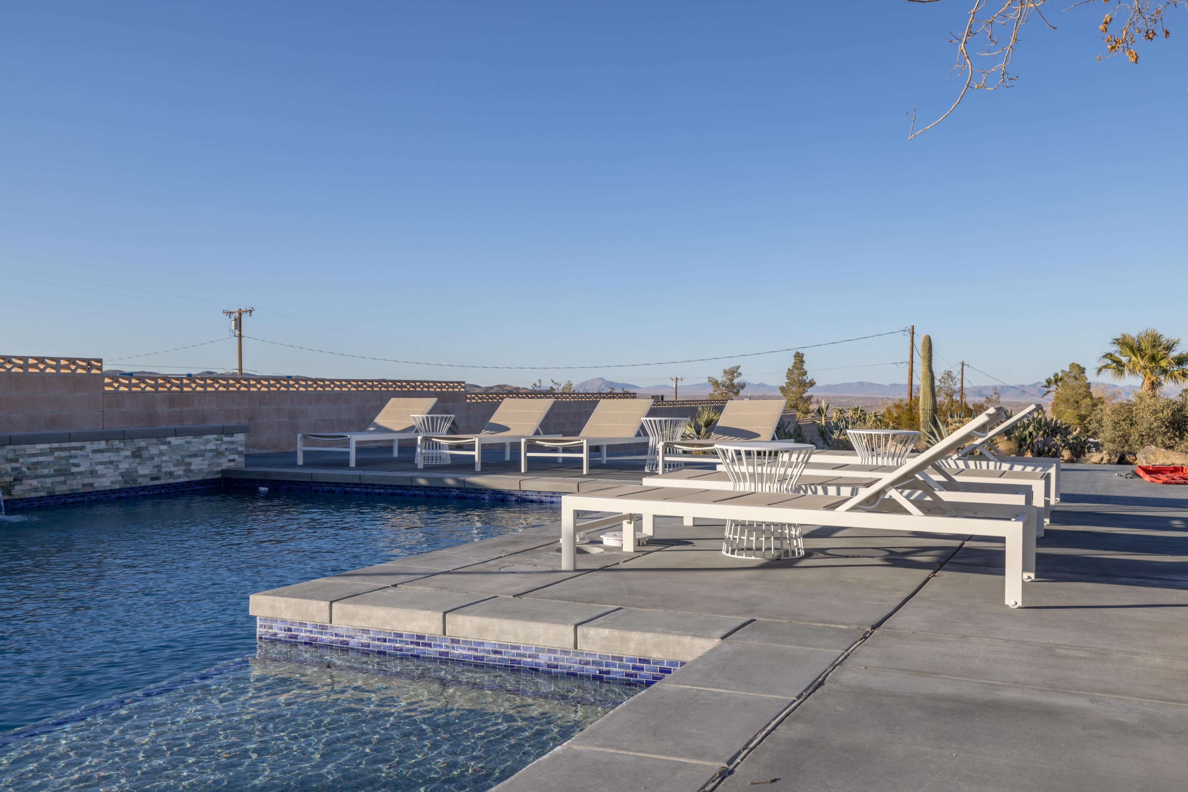 The image shows a modern swimming pool area with several white lounge chairs arranged around it, surrounded by sparse desert landscaping.