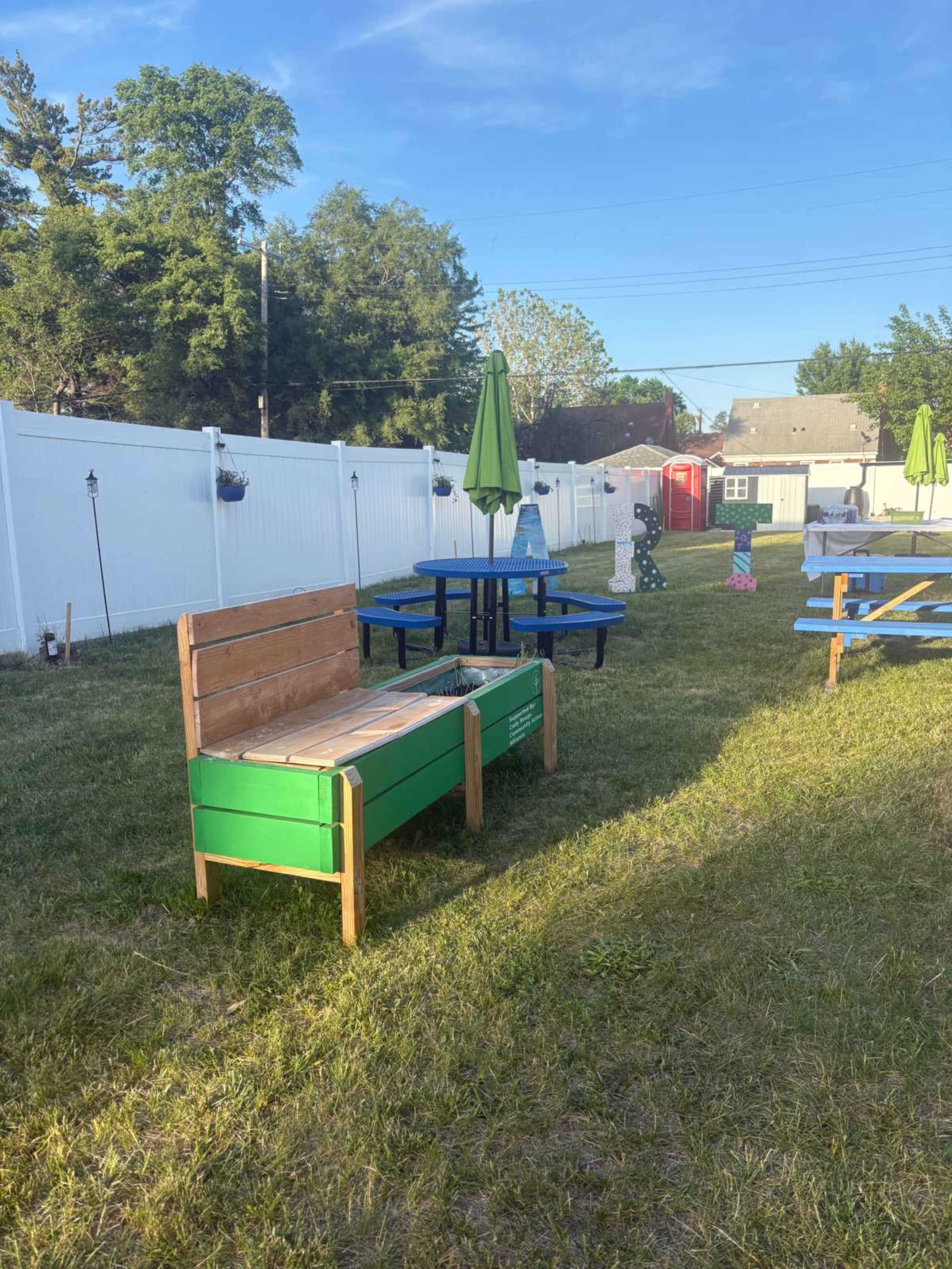 The image shows a grassy area with several outdoor picnic tables, a green storage bench, and a white fence in the background.