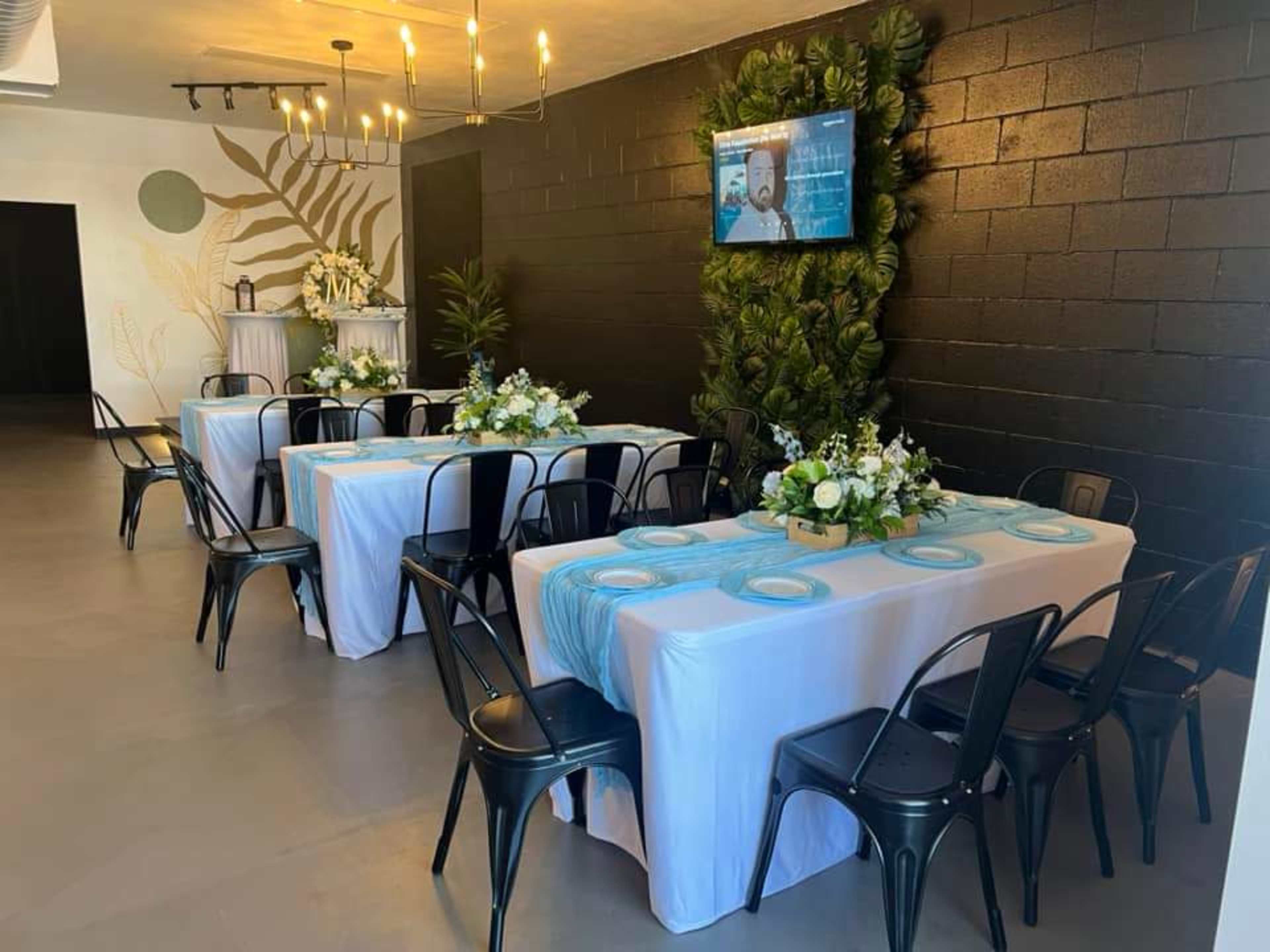 The image shows a decorated indoor venue with two rows of tables set for an event, featuring blue table runners and floral centerpieces, against a backdrop of greenery and a television screen on a black wall.