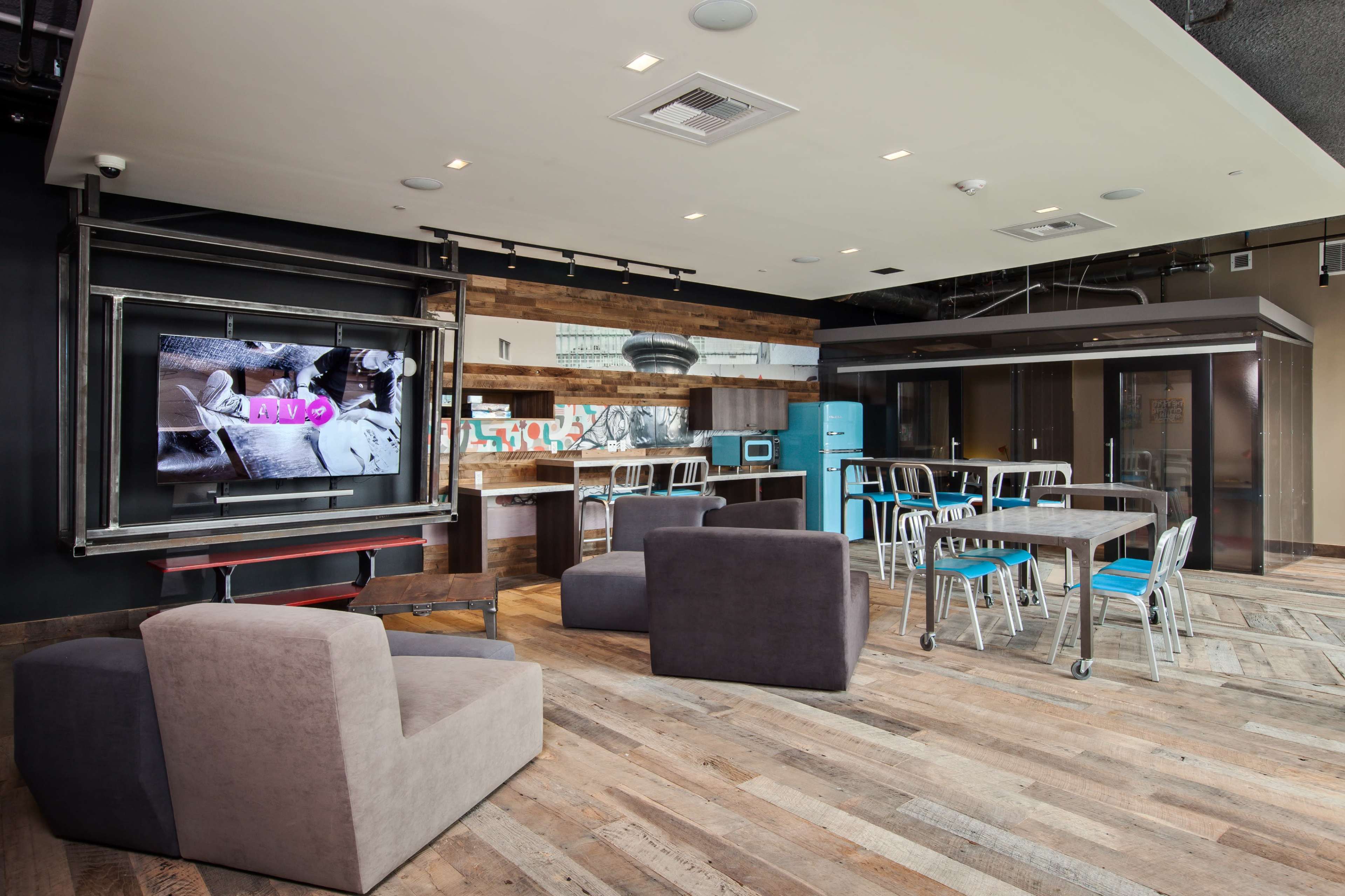 A modern communal space featuring a kitchen area, a large television, and various seating options arranged around a wooden floor.