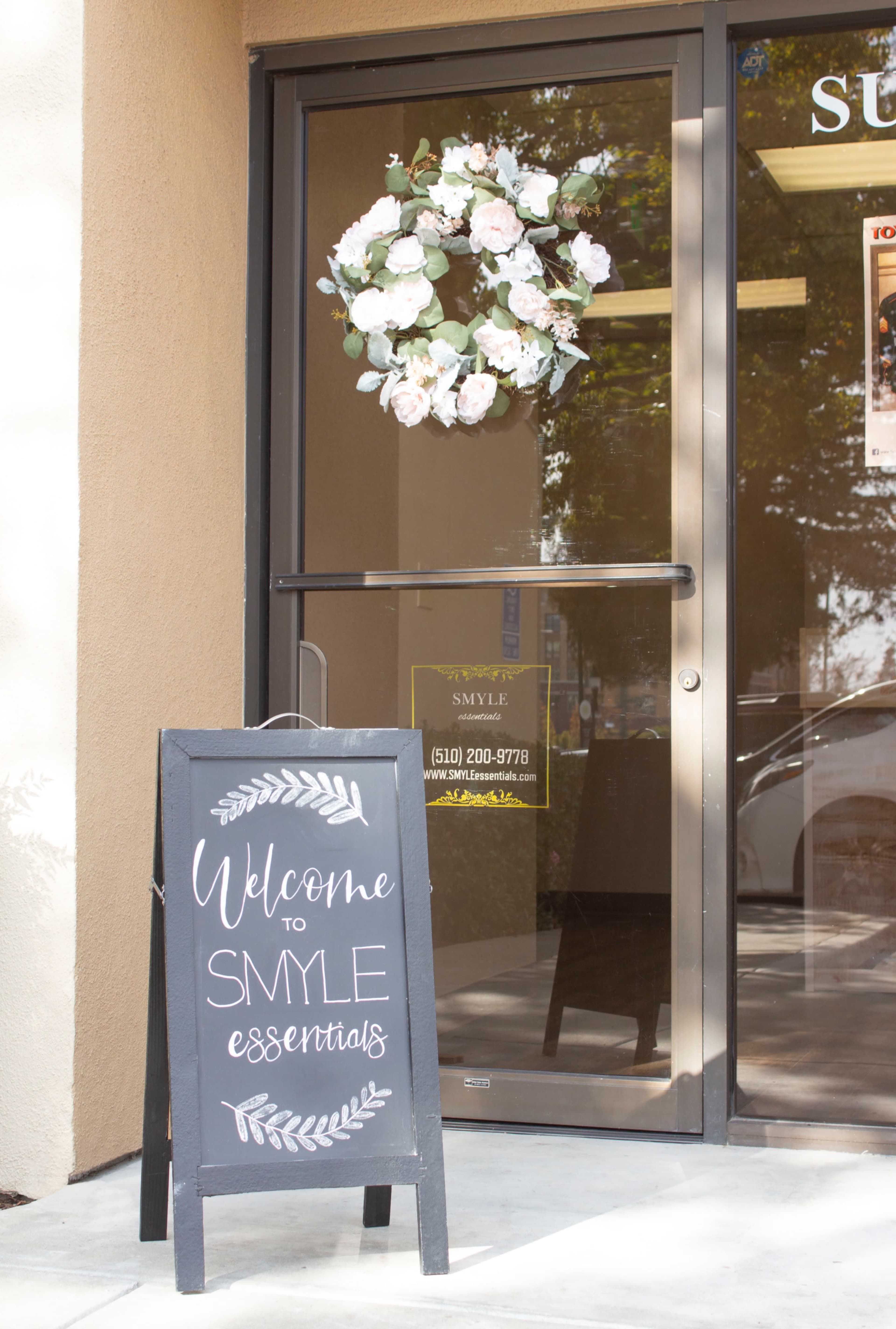 A sign reading "Welcome to SMYLE essentials" stands in front of a glass door adorned with a floral wreath.