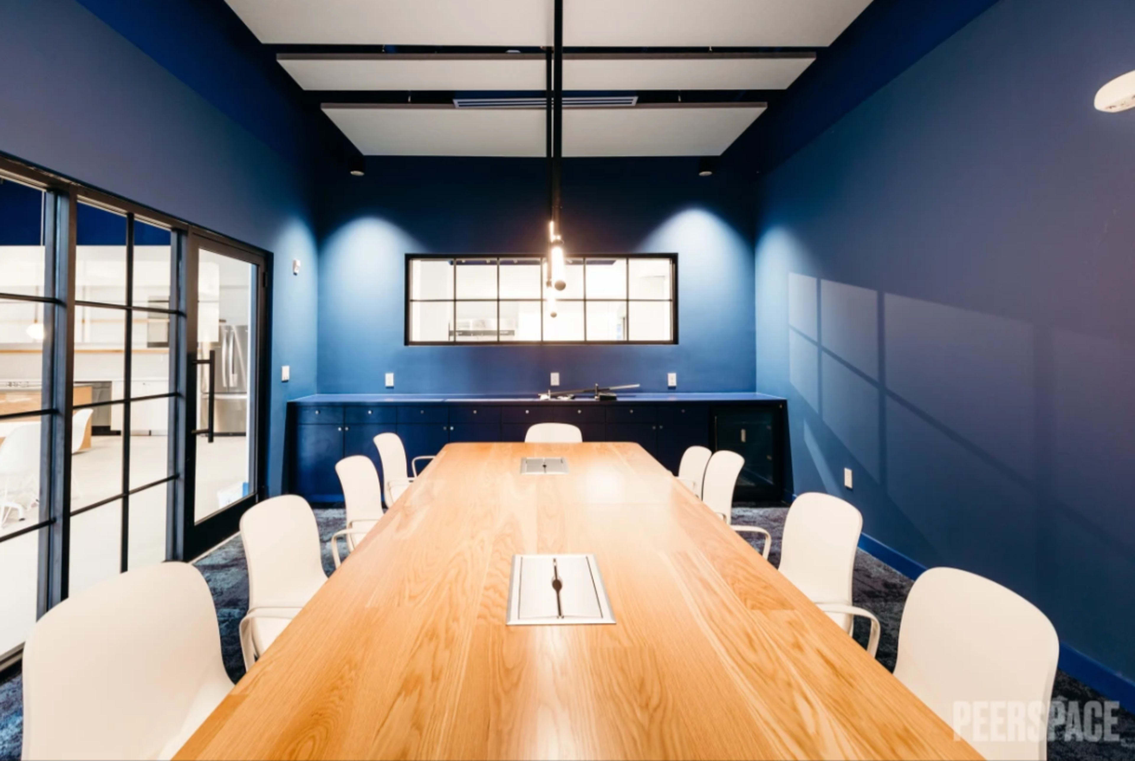 A modern conference room with a long wooden table surrounded by white chairs, set against deep blue walls and featuring large windows.