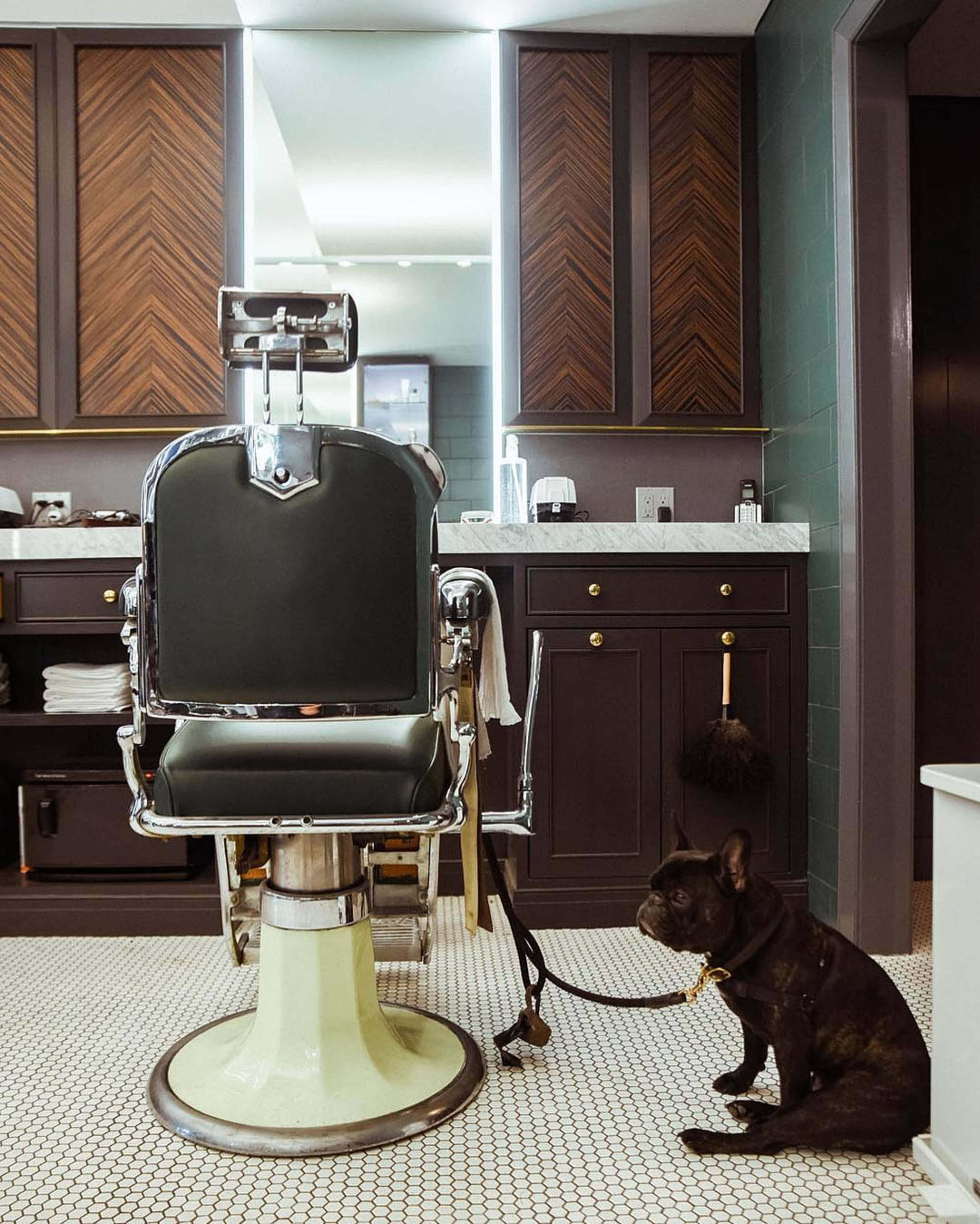 A classic barber chair stands in front of a mirror, with a French Bulldog sitting on the tiled floor beside it.