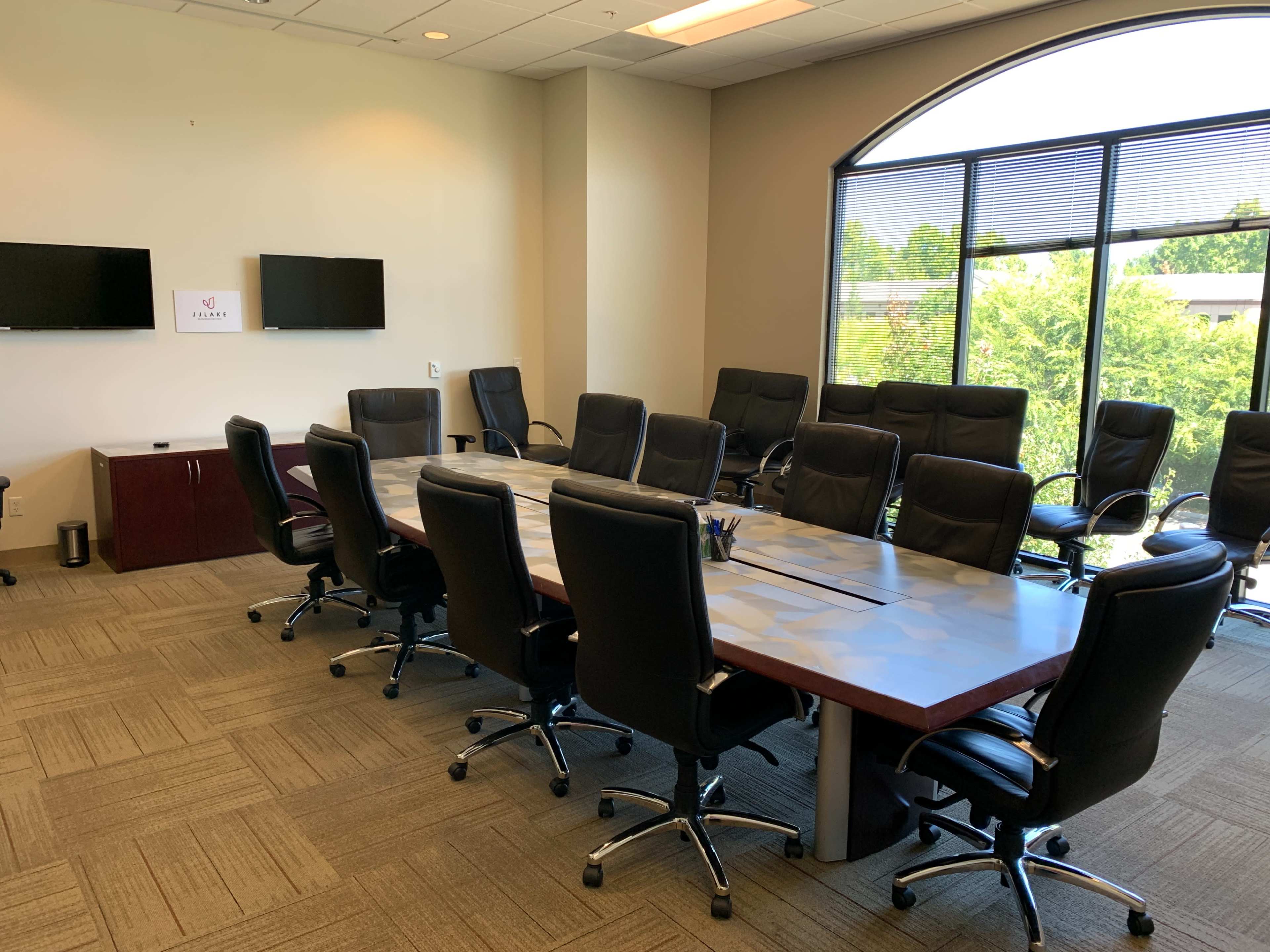 A large, modern conference room with a rectangular table surrounded by black ergonomic chairs, flanked by two wall-mounted screens.