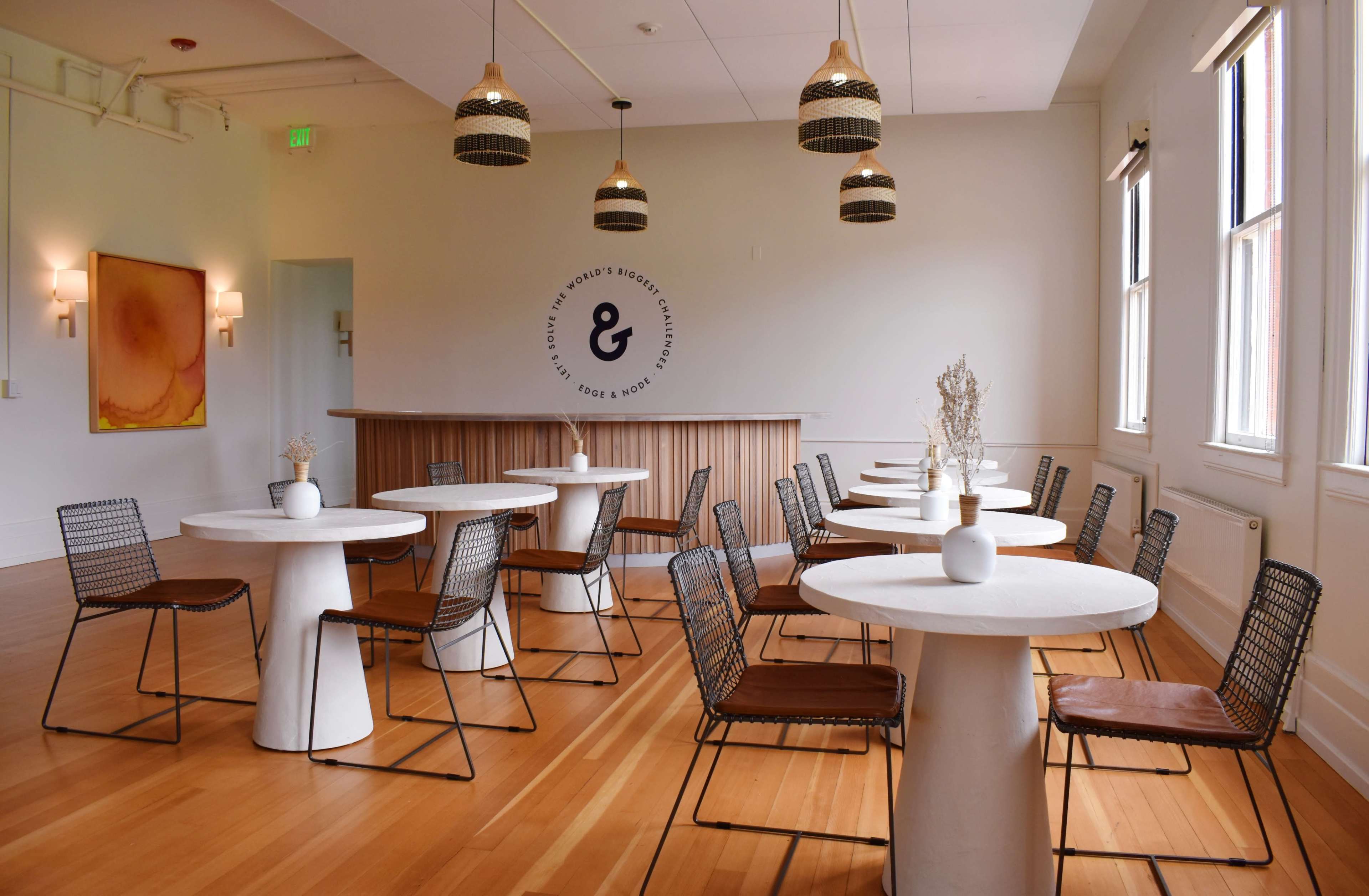A modern cafe interior featuring white round tables and black wire chairs arranged neatly on a wooden floor, with pendant lights hanging above.