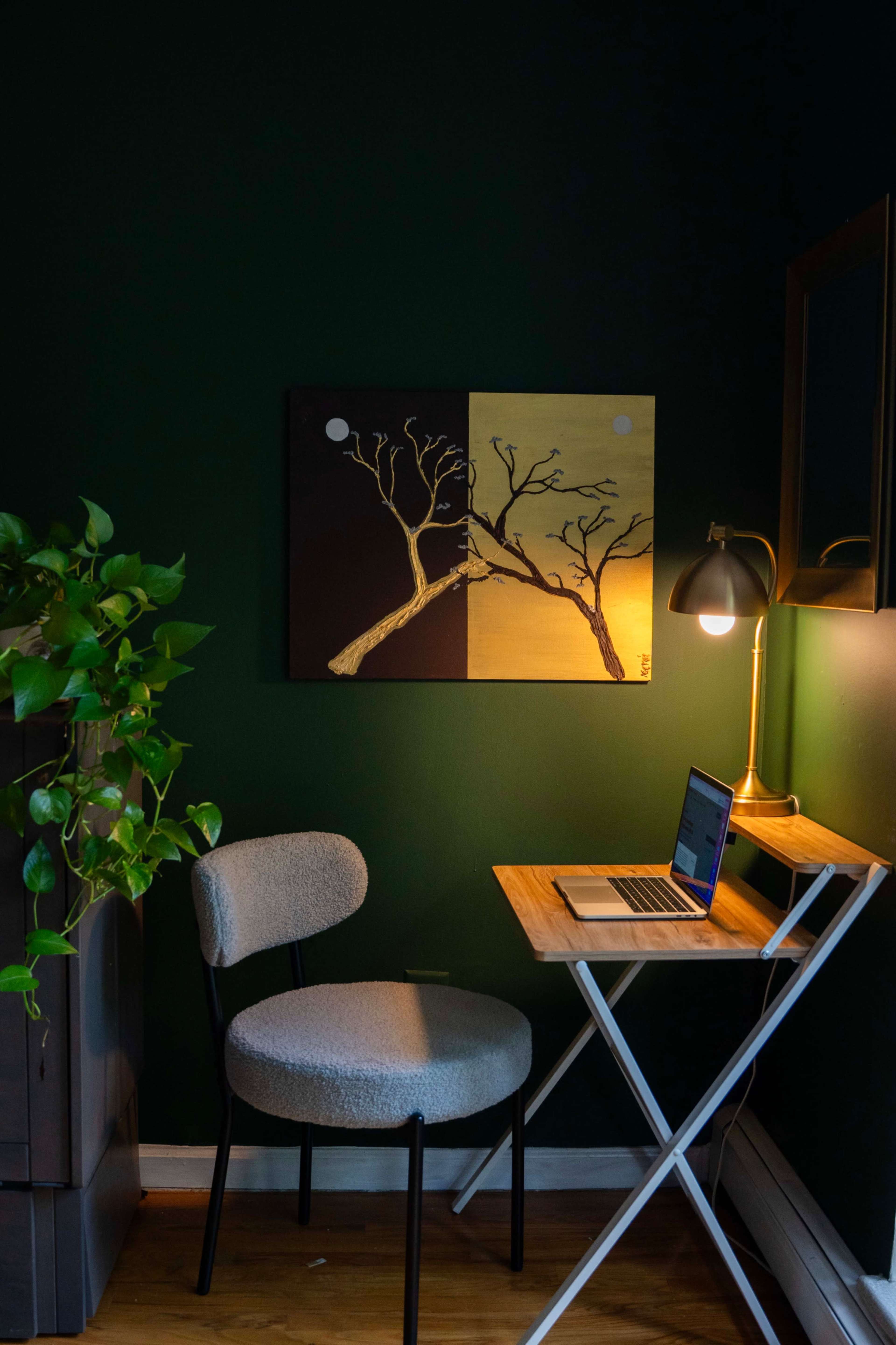 A small workspace features a light wooden table with a laptop, a gray chair, a potted plant, and a wall-mounted mirror against a dark green backdrop, with an abstract painting above the table.