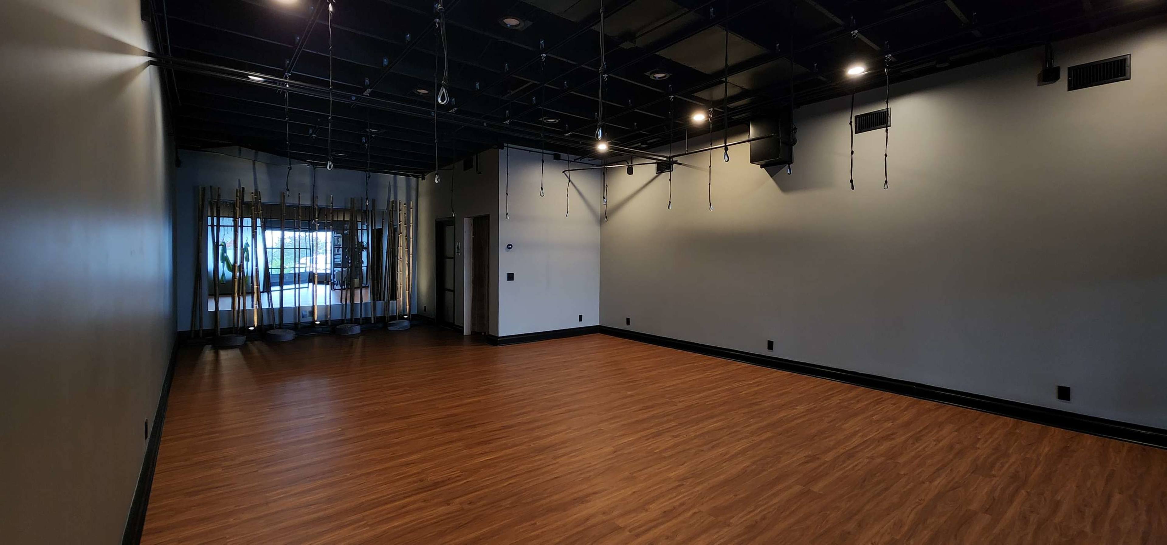 A spacious, empty room with wooden flooring and black ceiling tiles, featuring a few wall-mounted mirrors on one side.