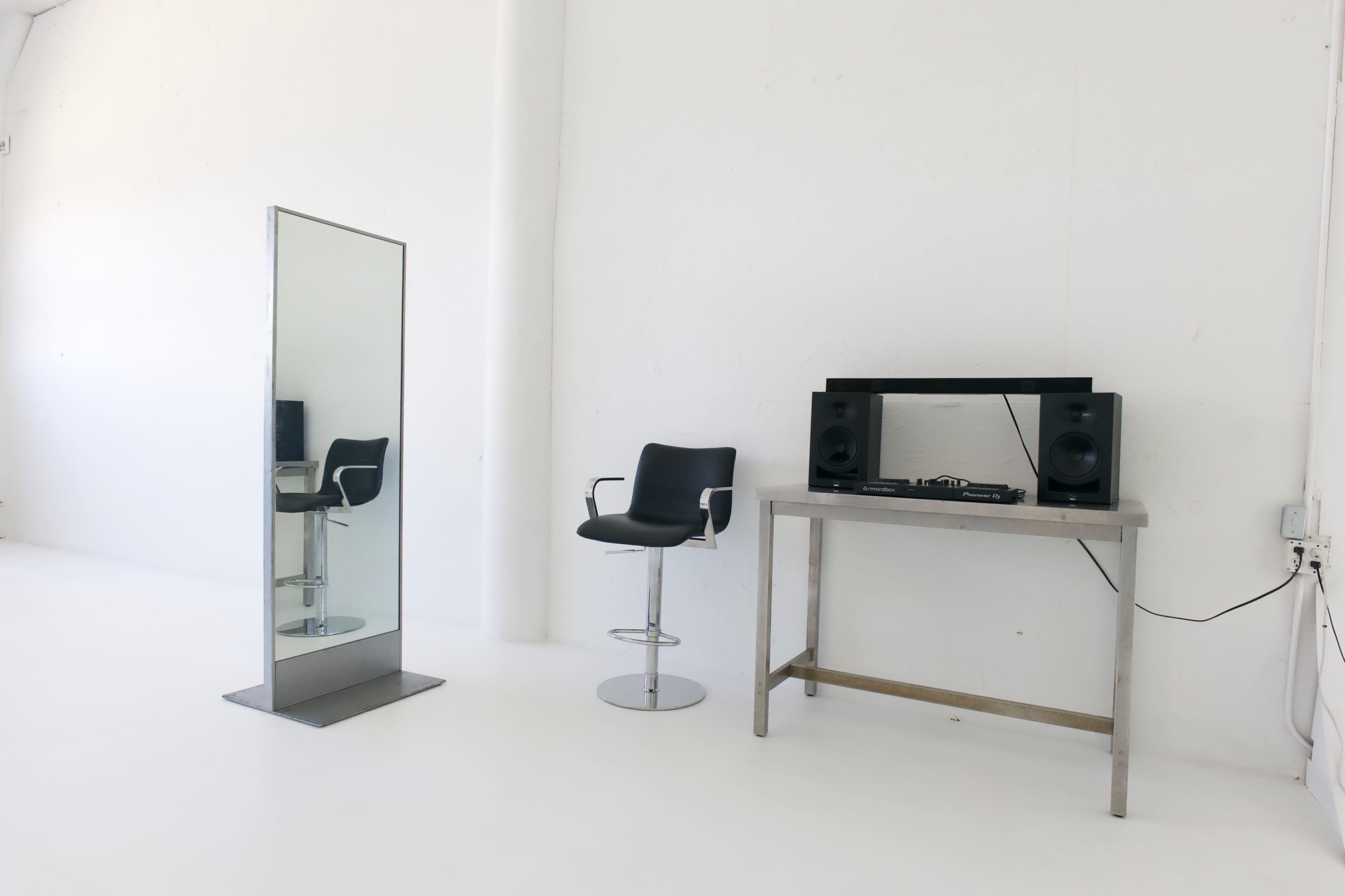 The image shows a minimalist room with a full-length mirror on the left, a black barstool, and a silver table with audio equipment on the right.