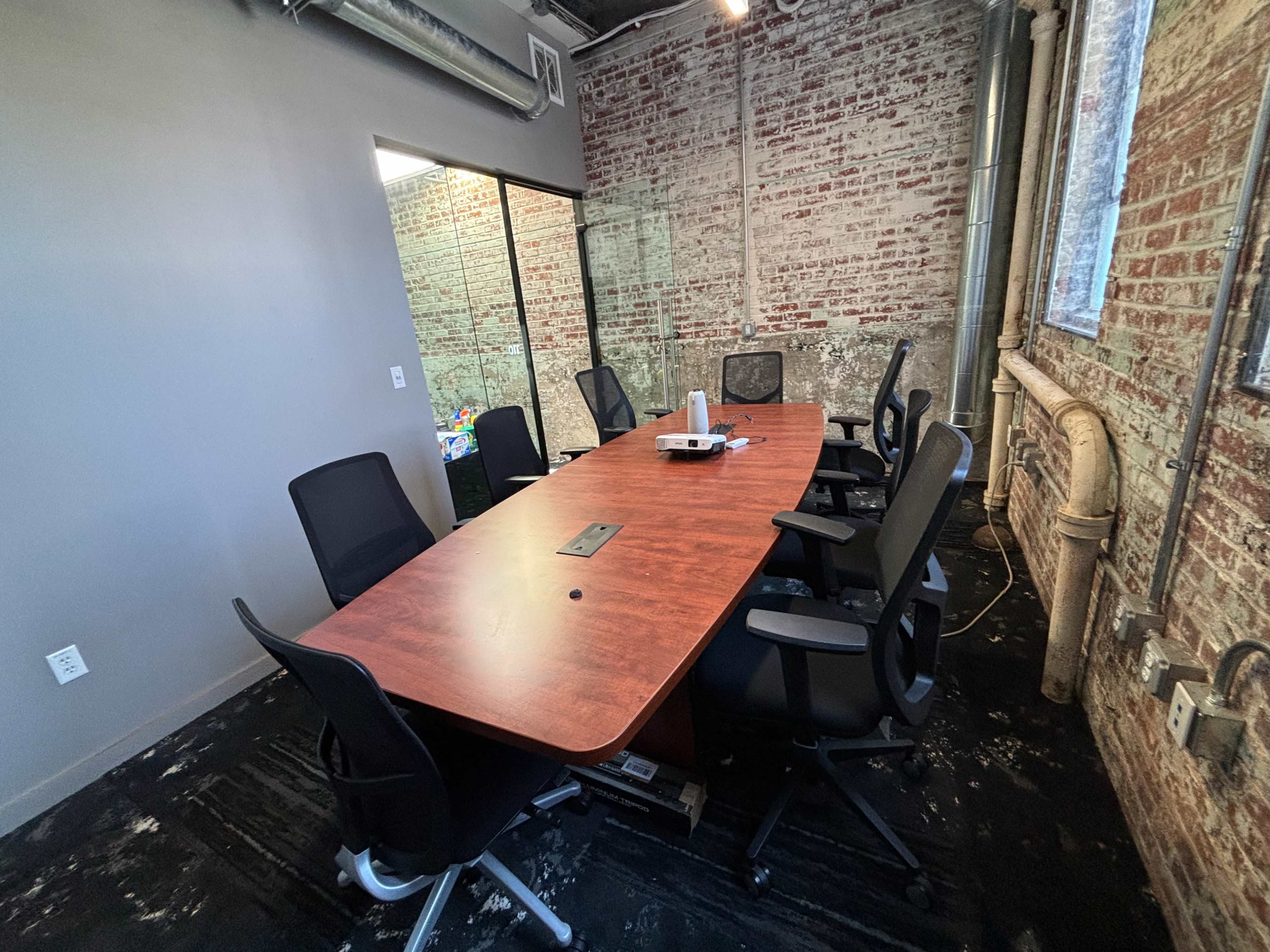 A conference room features a long wooden table surrounded by black chairs, with exposed brick walls and a glass-walled meeting area.