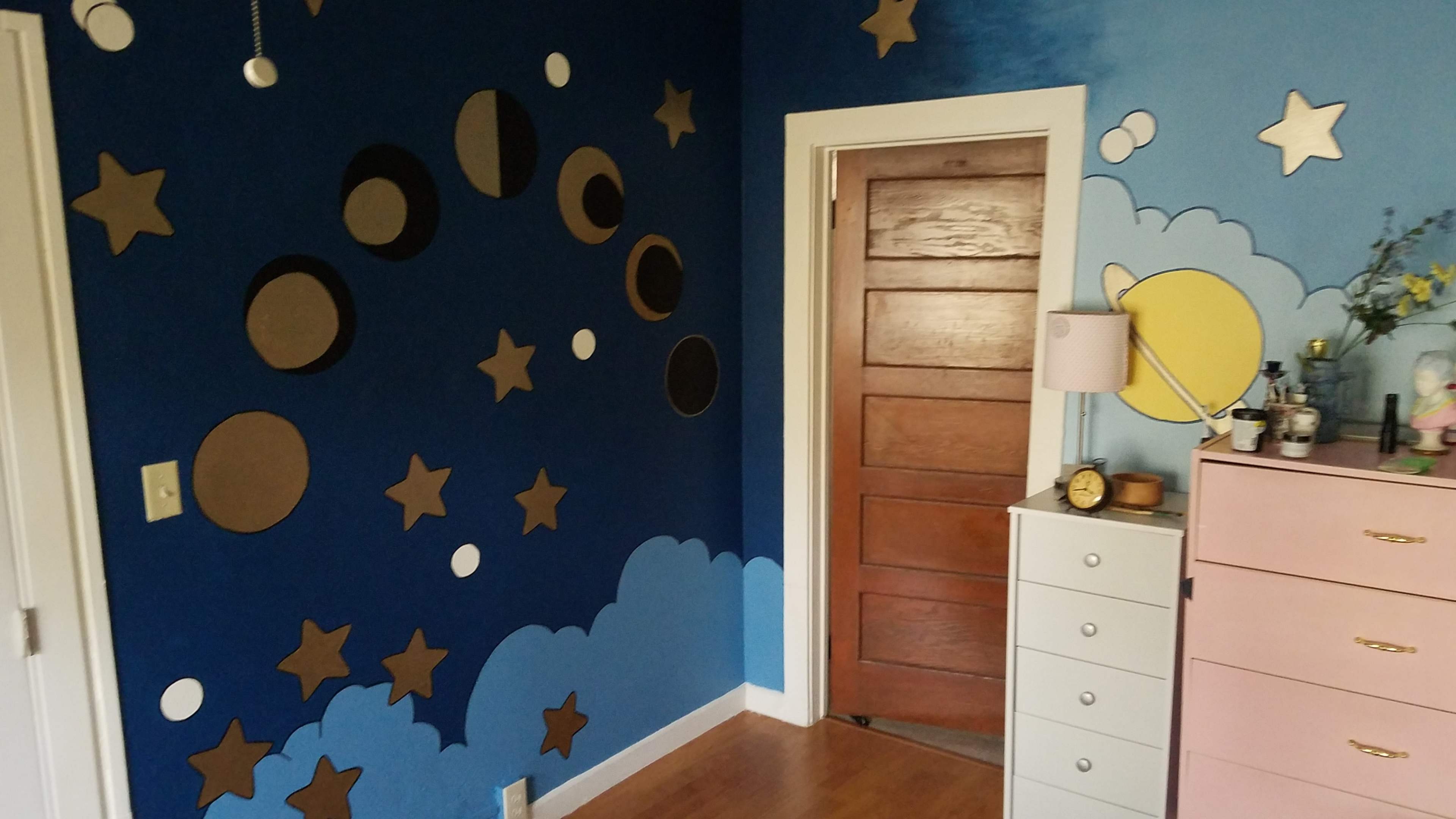 The image shows a room with a painted blue wall featuring clouds and stars, and a wooden door leading to another area.