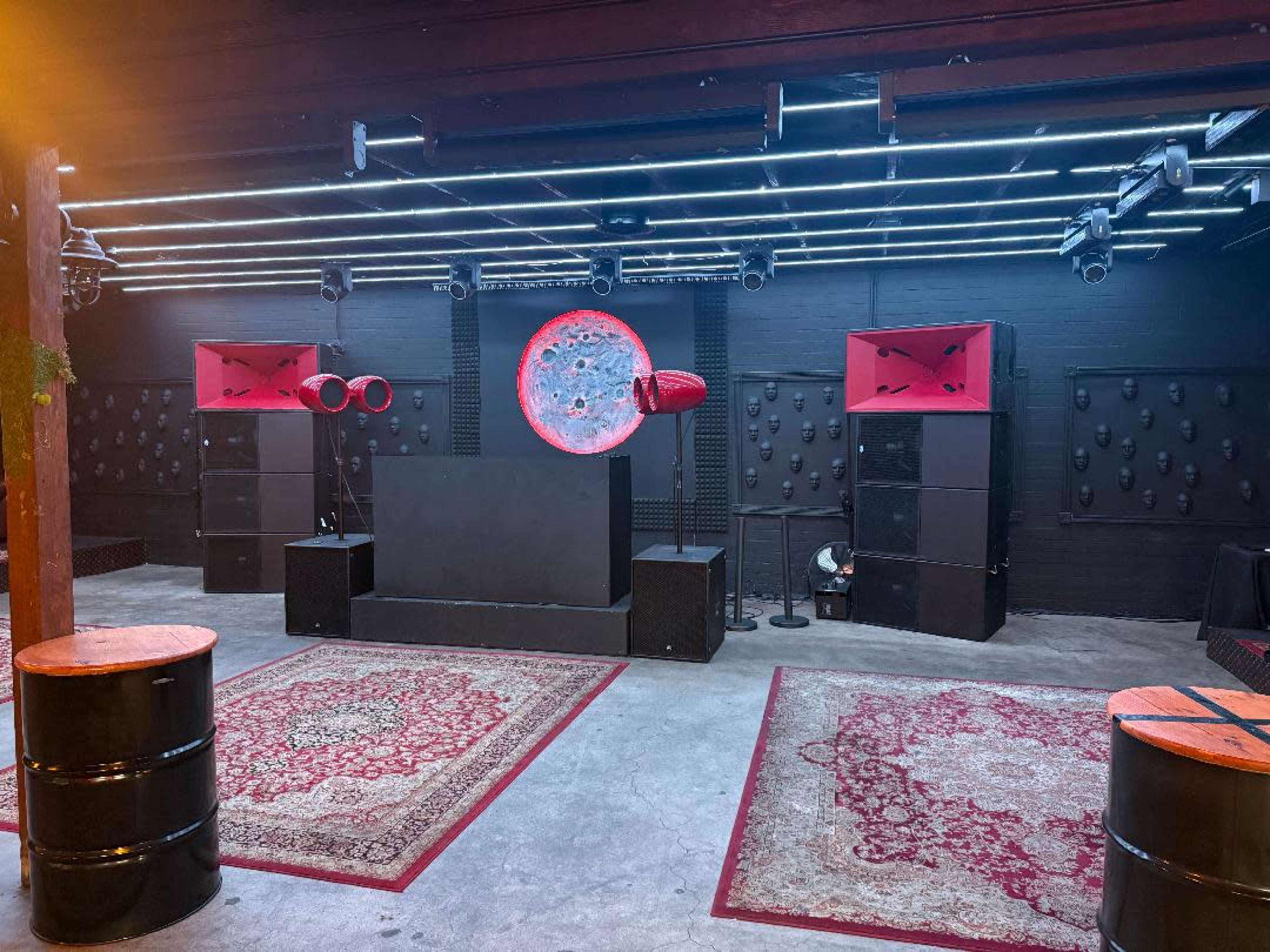 The image shows a music venue setup with a stage, large speakers, and patterned rugs on a concrete floor.