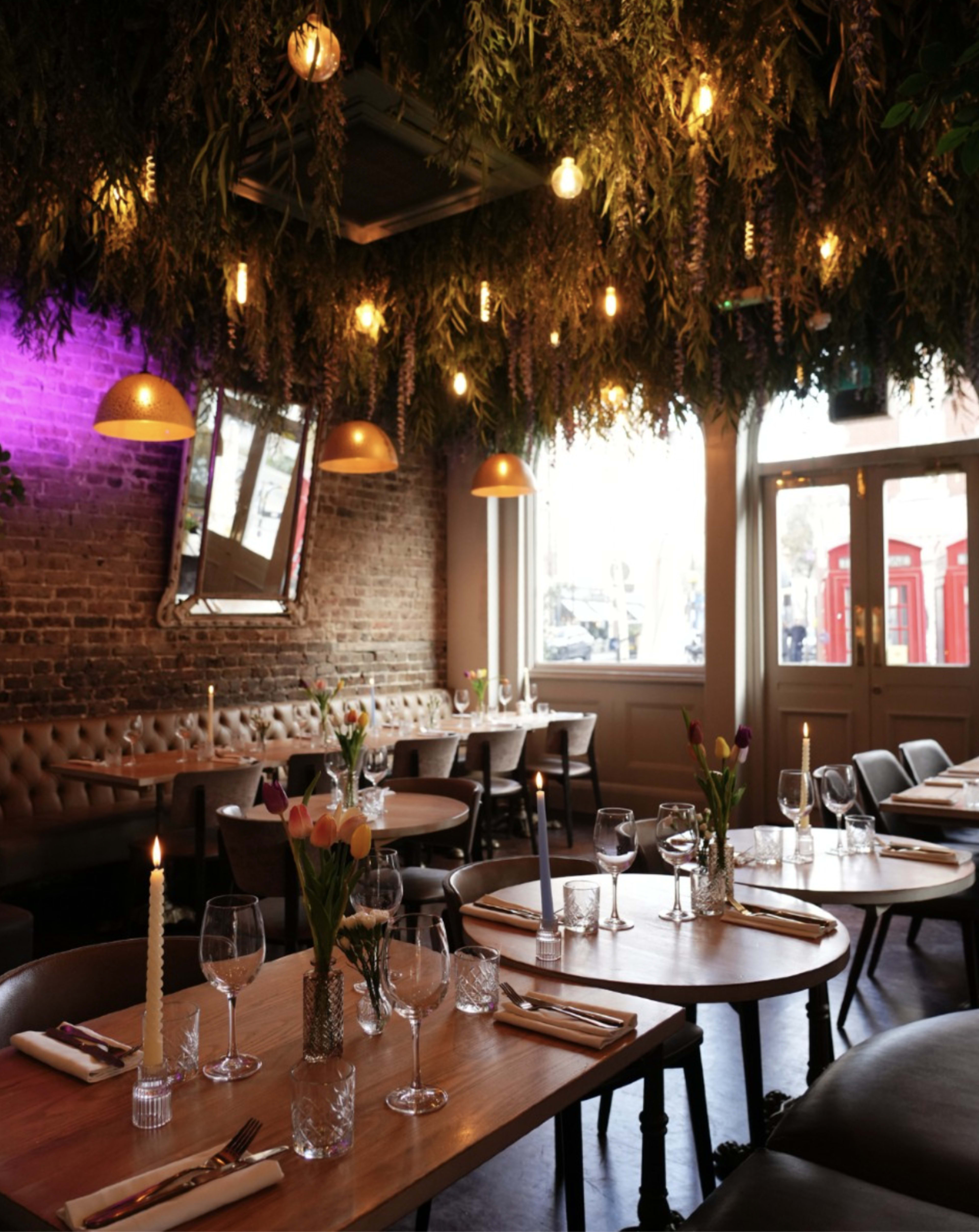 The image shows a cozy restaurant interior with tables set for dining, adorned with fresh flowers, candles, and a ceiling decorated with hanging greenery and lights.