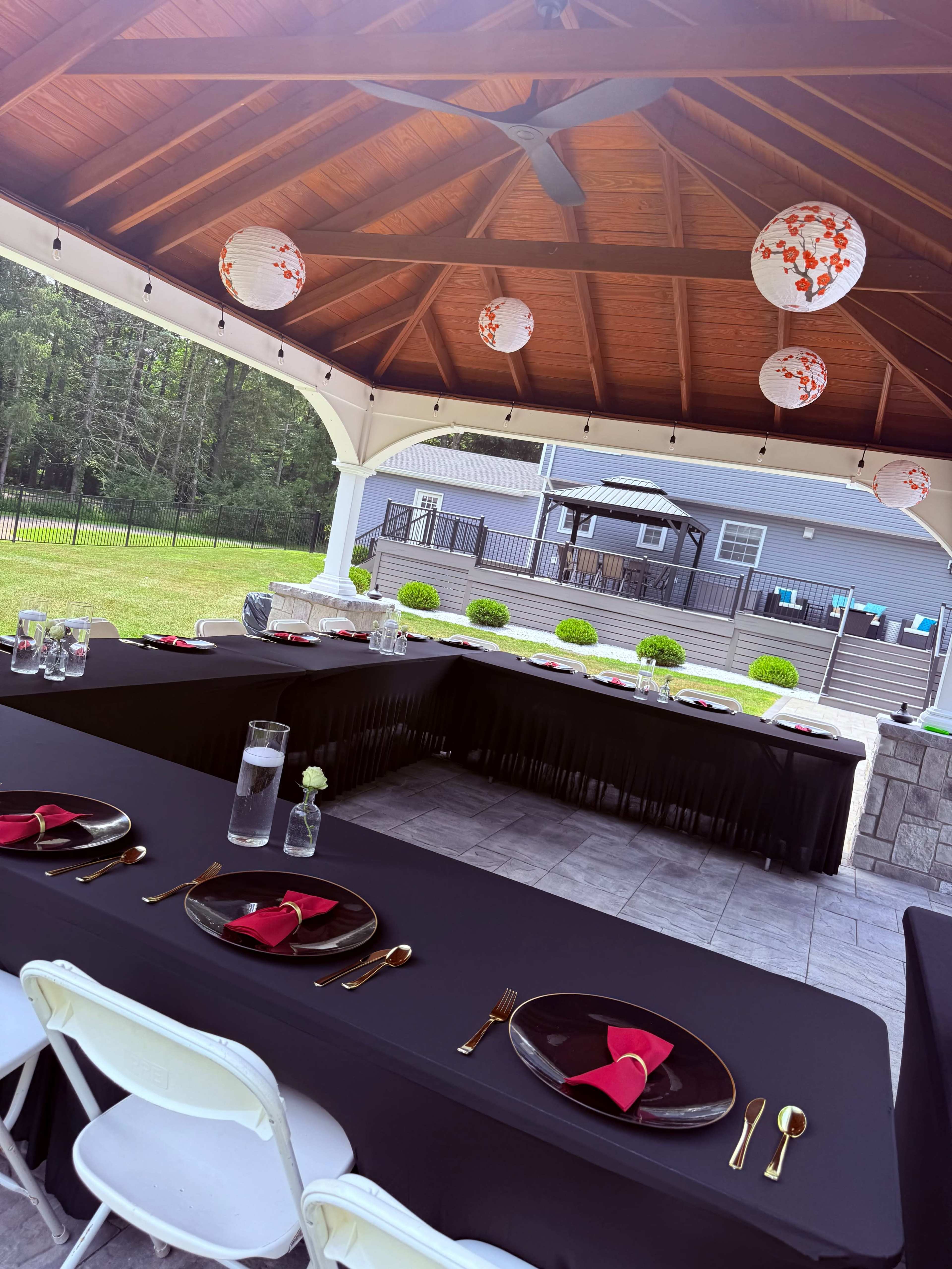 A covered outdoor dining setup features tables with black linens, gold utensils, and red napkins, illuminated by decorative lanterns hanging from the ceiling.