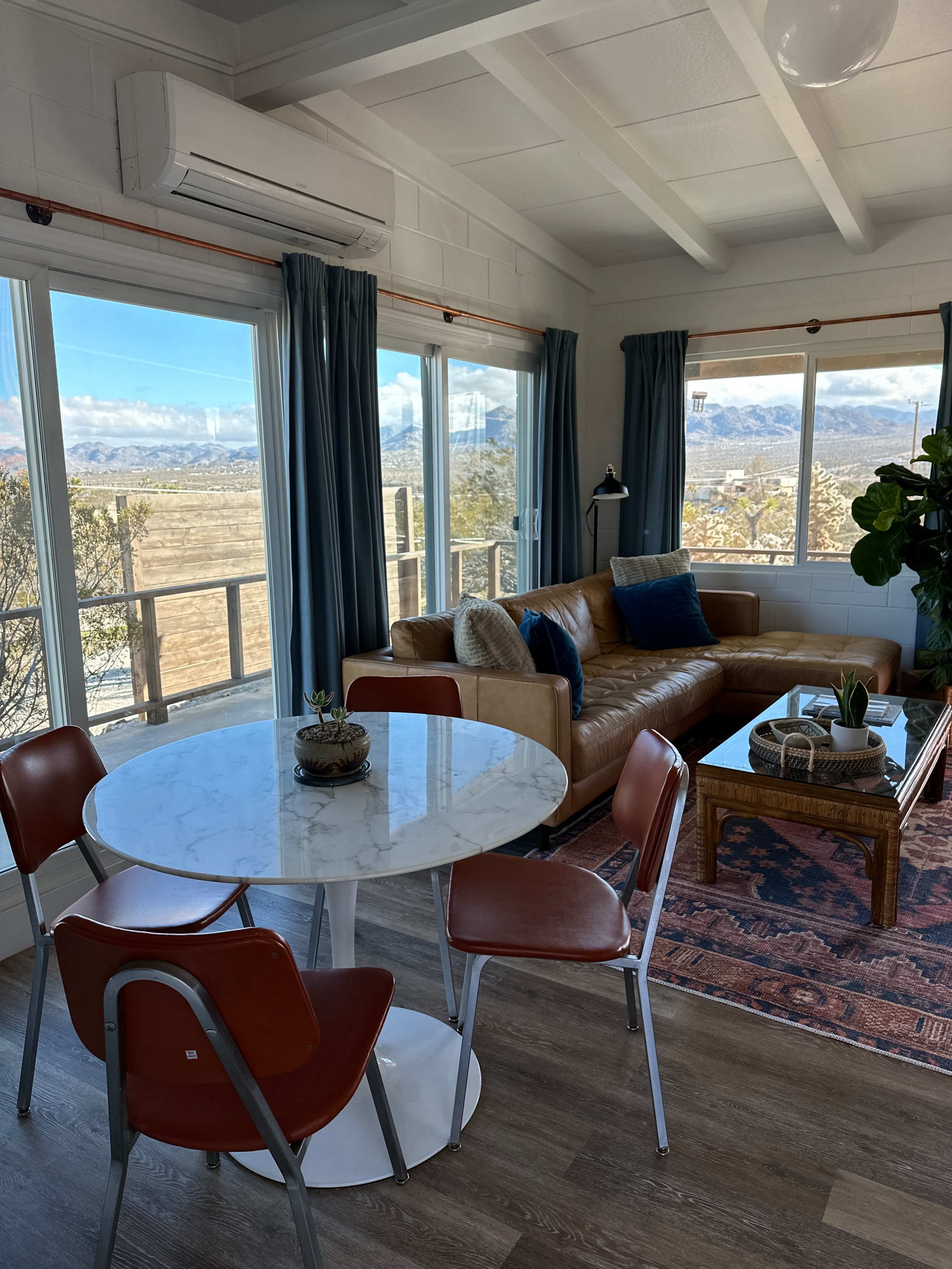 A living area with a circular marble dining table, a brown leather sofa, and large windows offering a view of a landscape with mountains in the background.