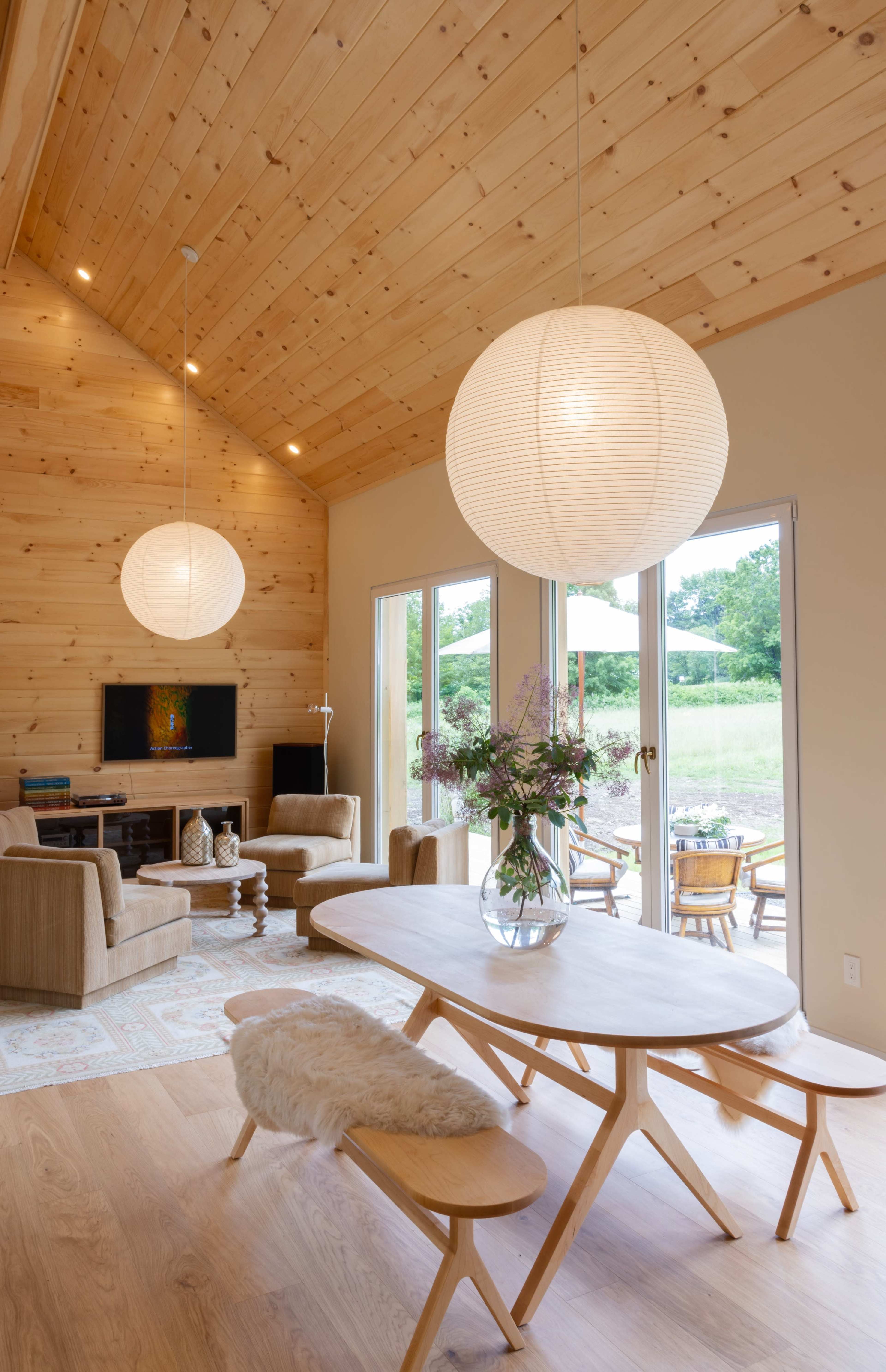 A modern living space with a wooden ceiling, a dining table surrounded by chairs, and large windows overlooking a green landscape.