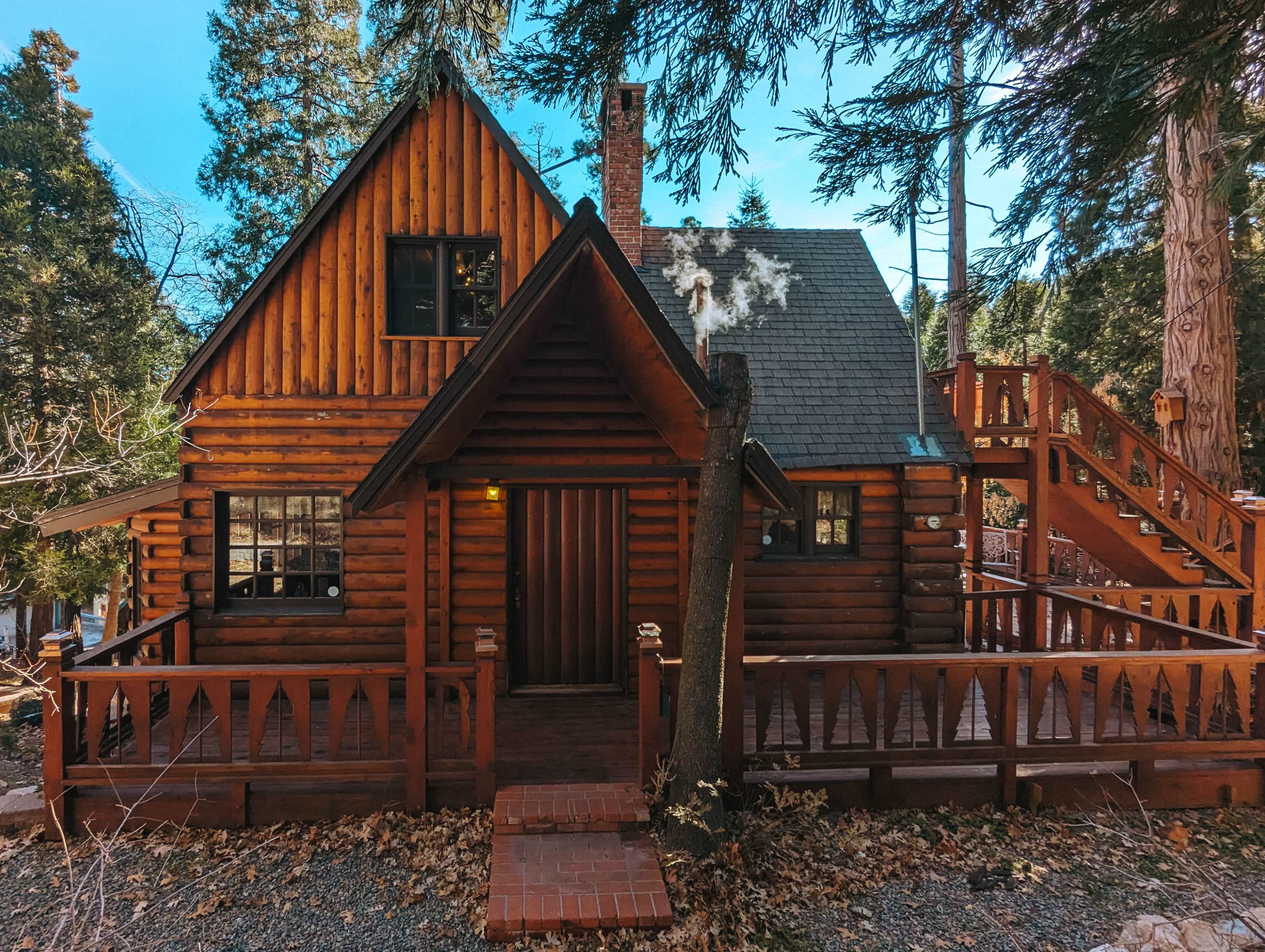 The image shows a wooden log cabin with a brick walkway and a spacious deck surrounded by trees.