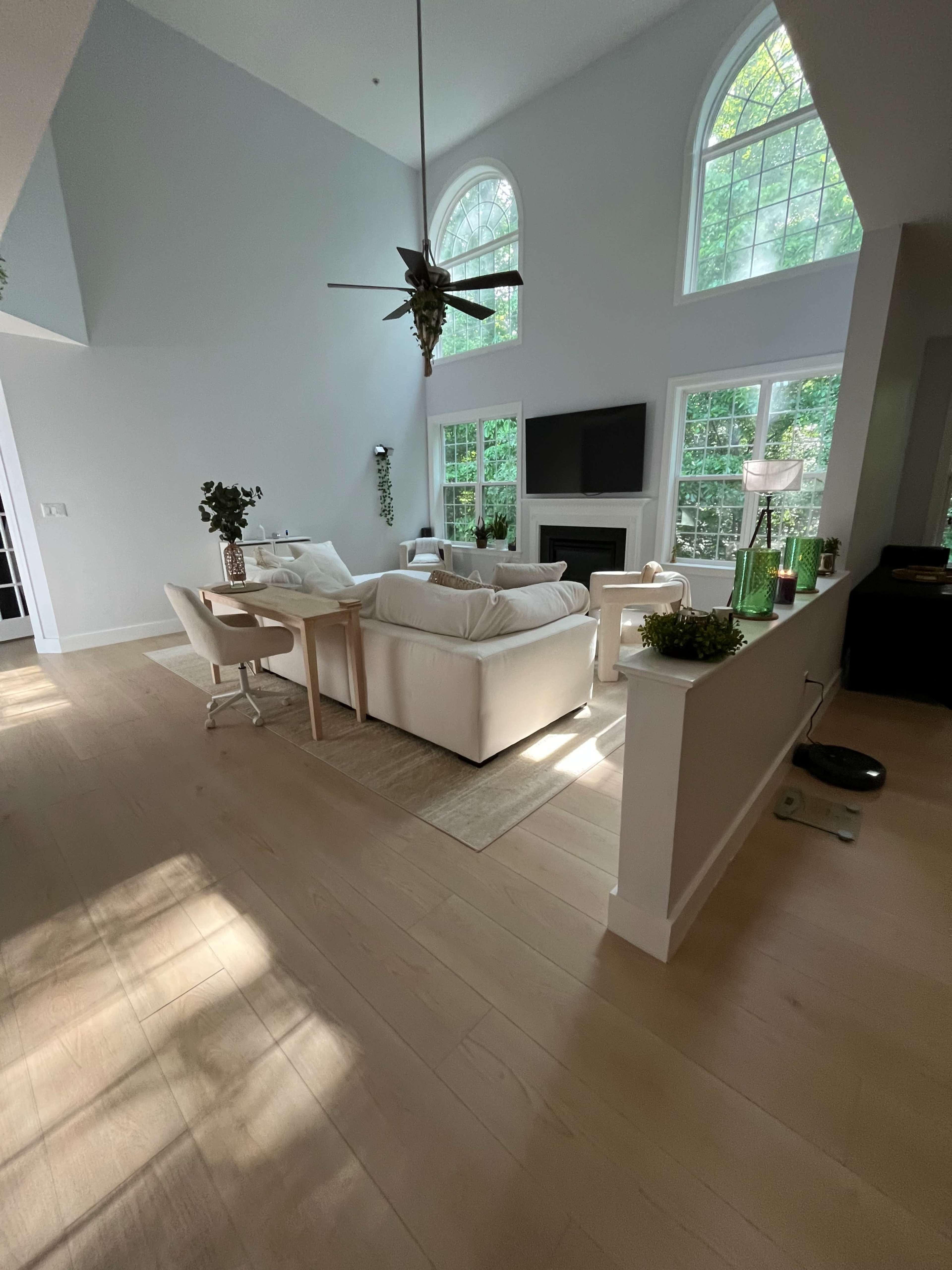 The image shows a bright and airy living room with large windows, a white sofa, and a central ceiling fan.