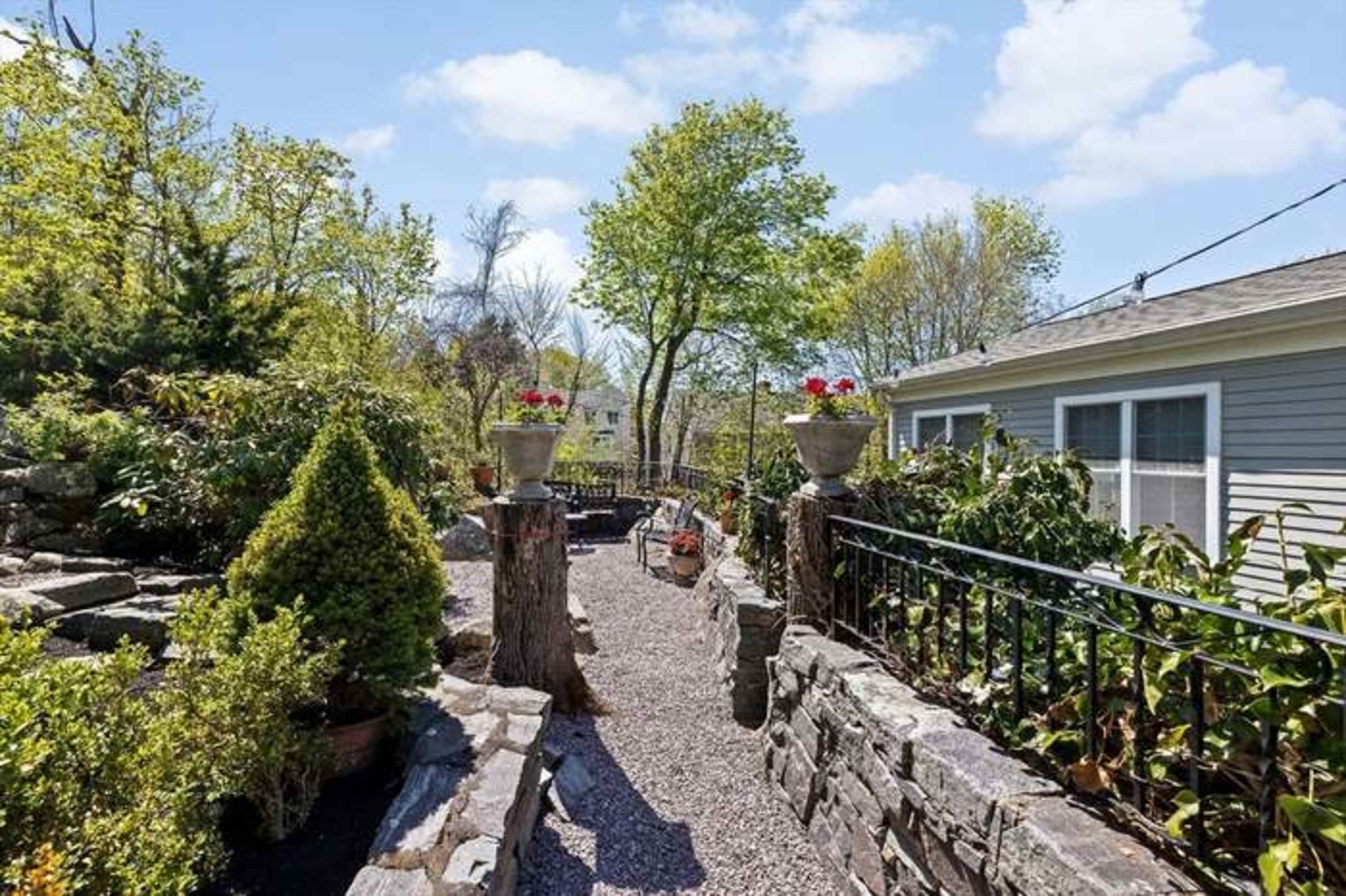 A narrow pathway lined with stone walls leads through a garden filled with potted plants and trees, with a house visible on the right.