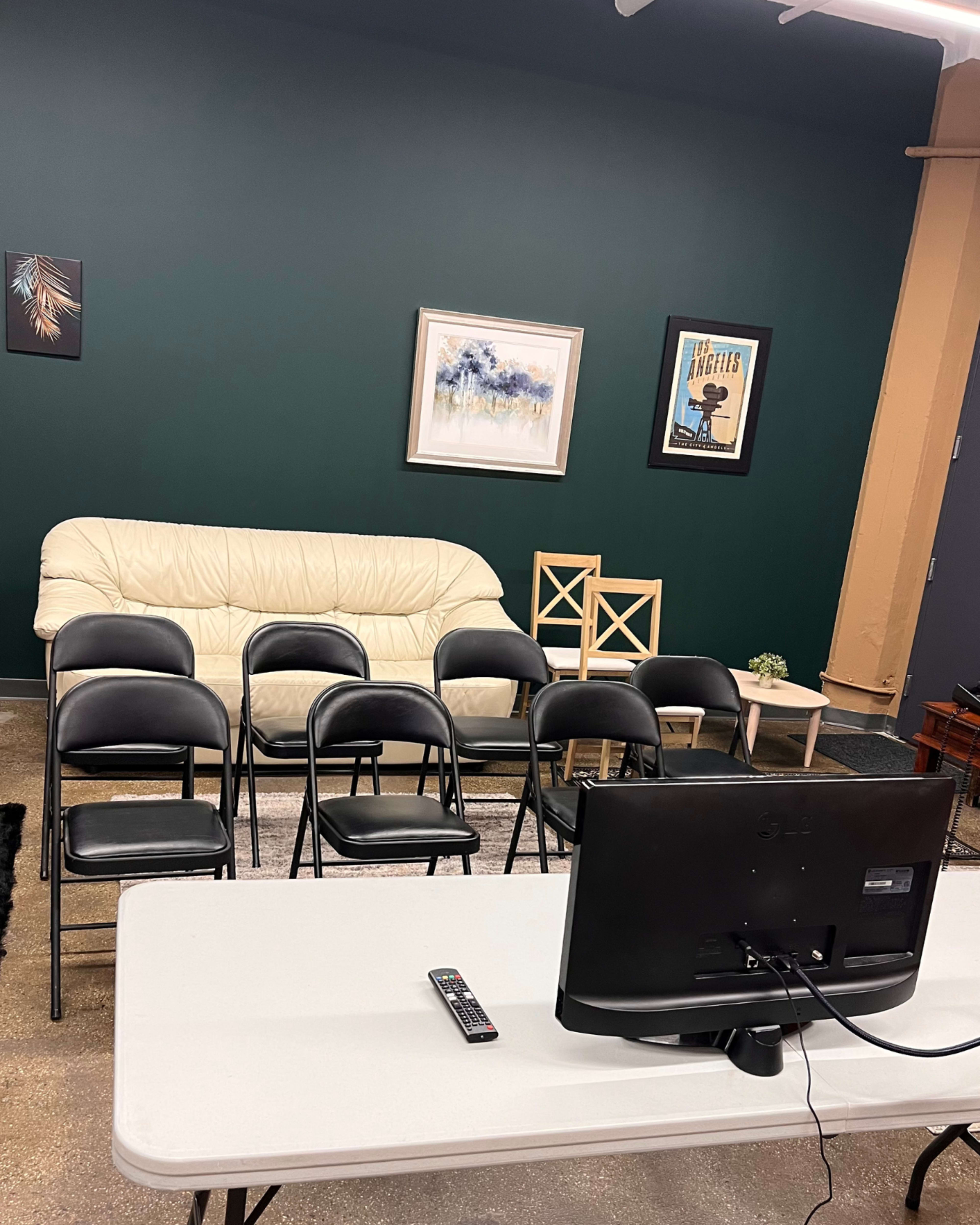 A room with a light-colored couch, several black chairs arranged in rows, a television on a table, and a framed picture on the wall.