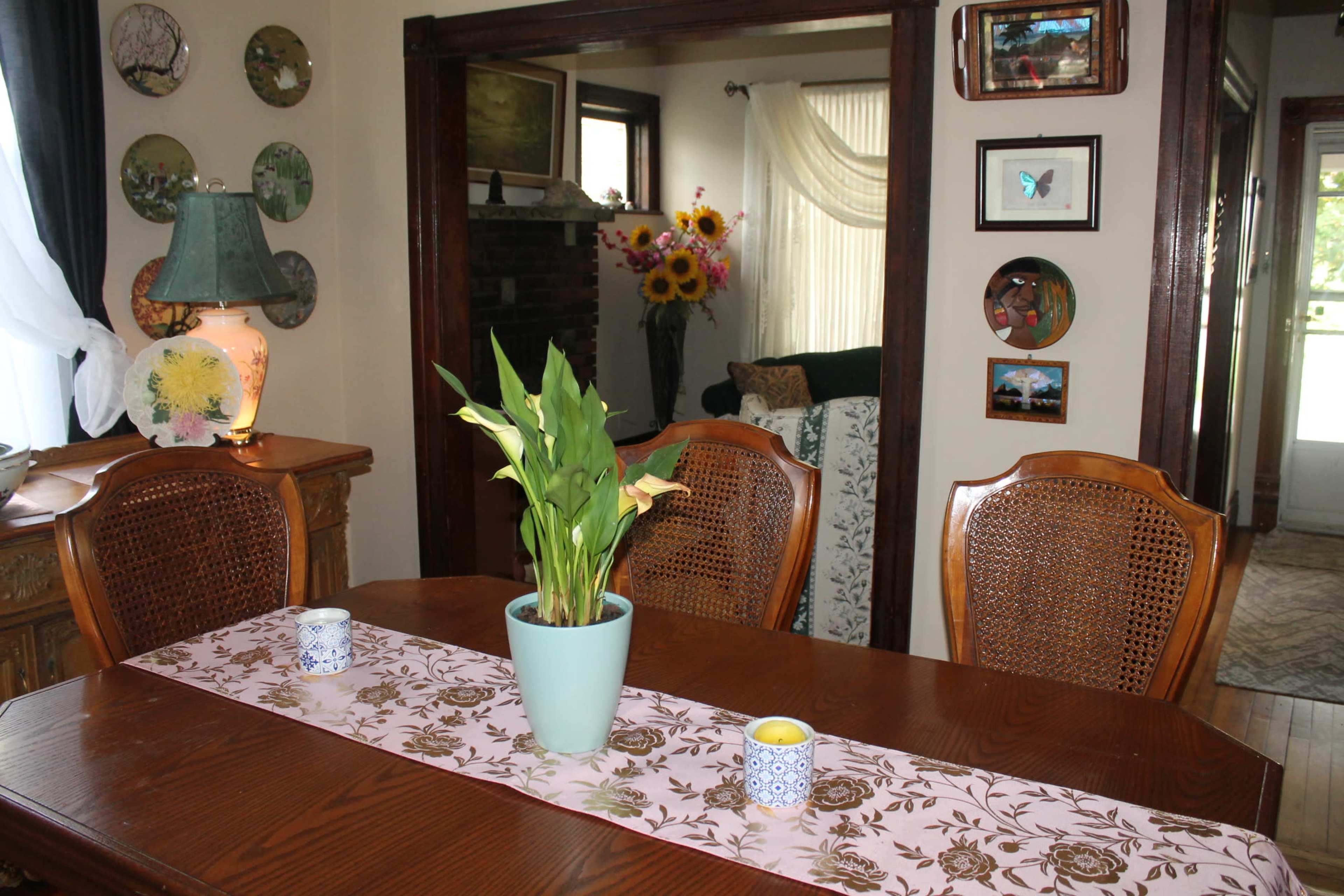 A wooden dining table with a pink table runner and a potted plant is set in a room adorned with art on the walls and a view into a living area.