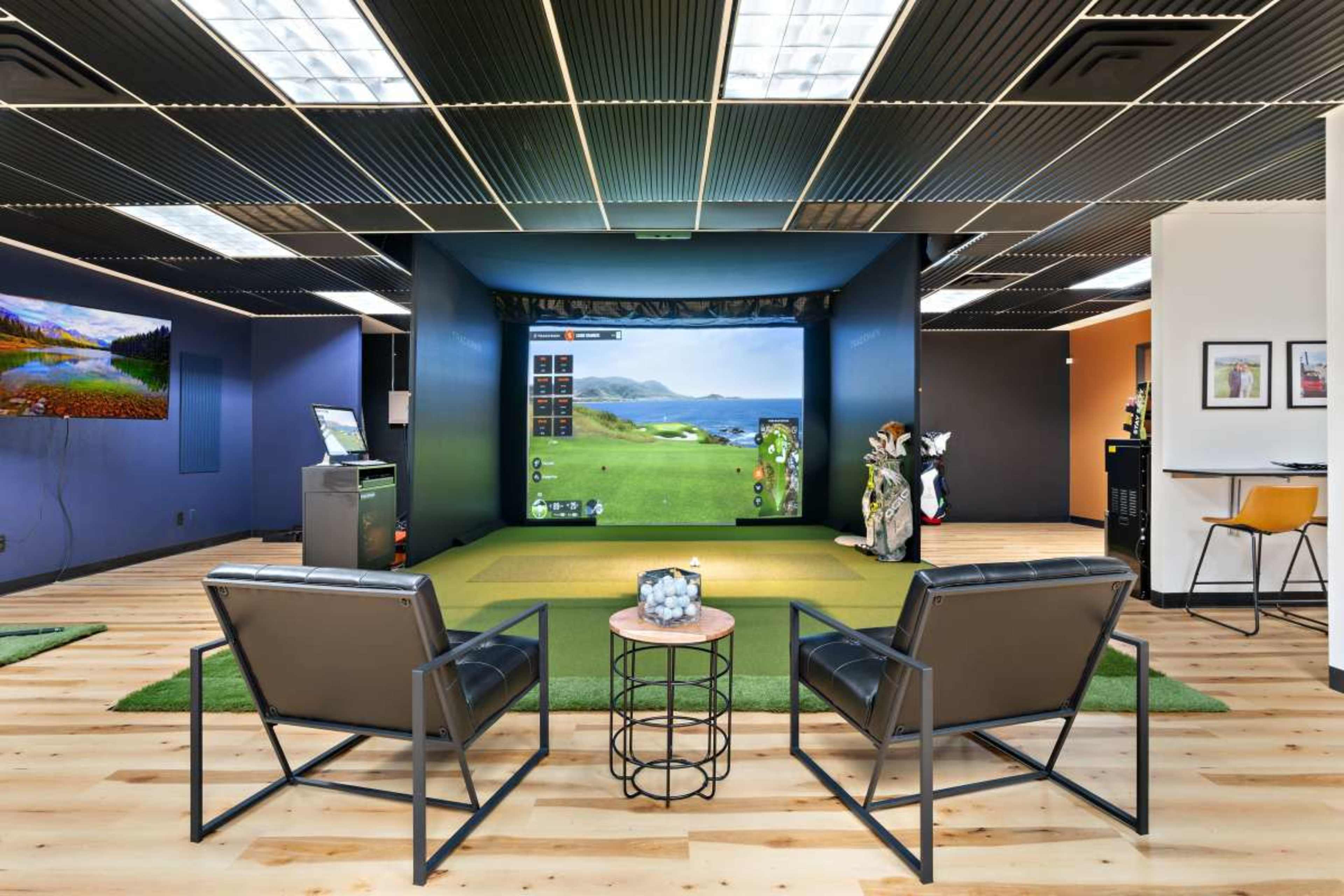 The image shows a modern indoor golf simulator setup with two chairs facing a large screen displaying a golf course.