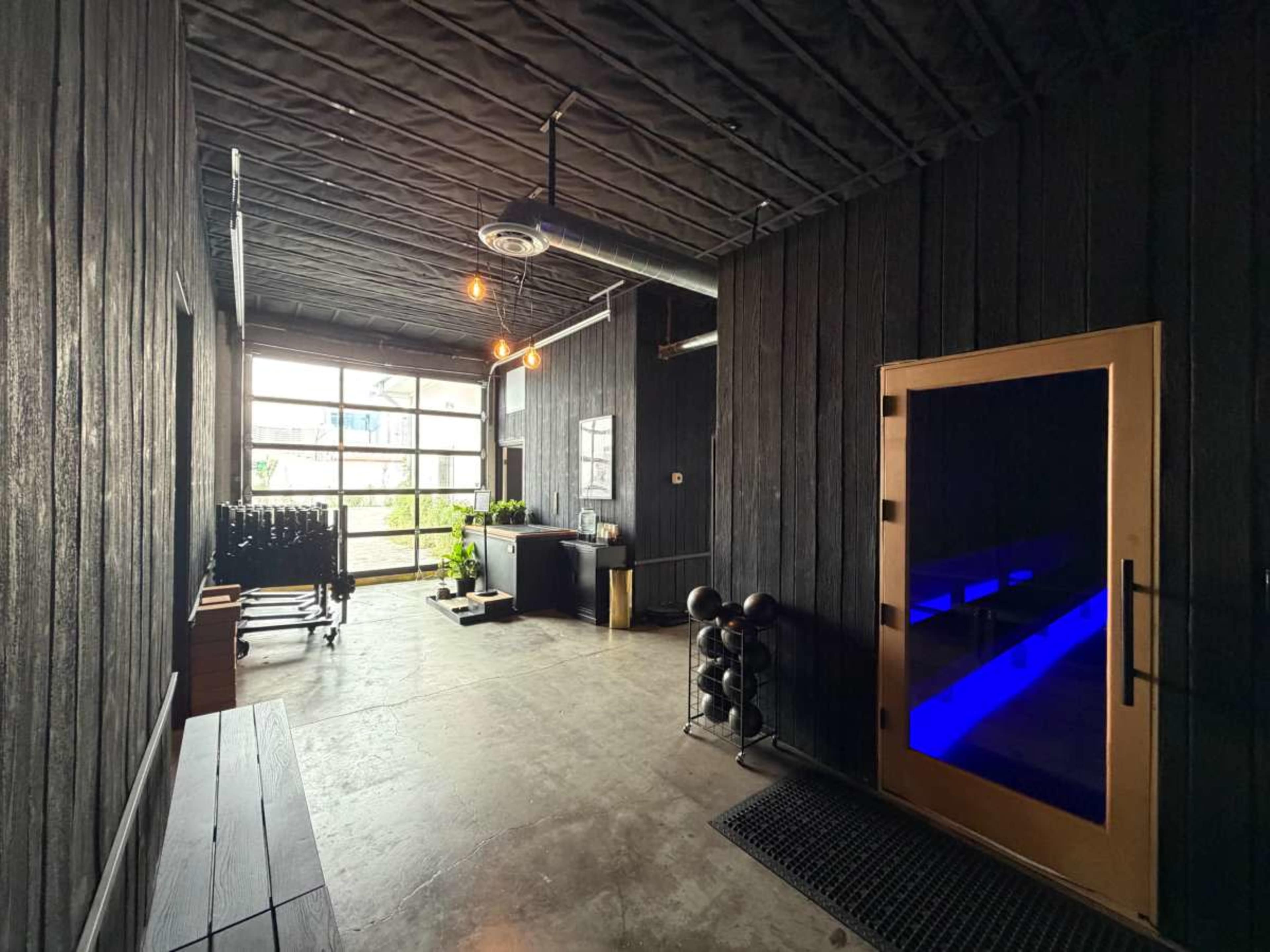 The image shows a modern hallway with dark wooden walls, a glass door at the end, and a workout area featuring weights and plants.