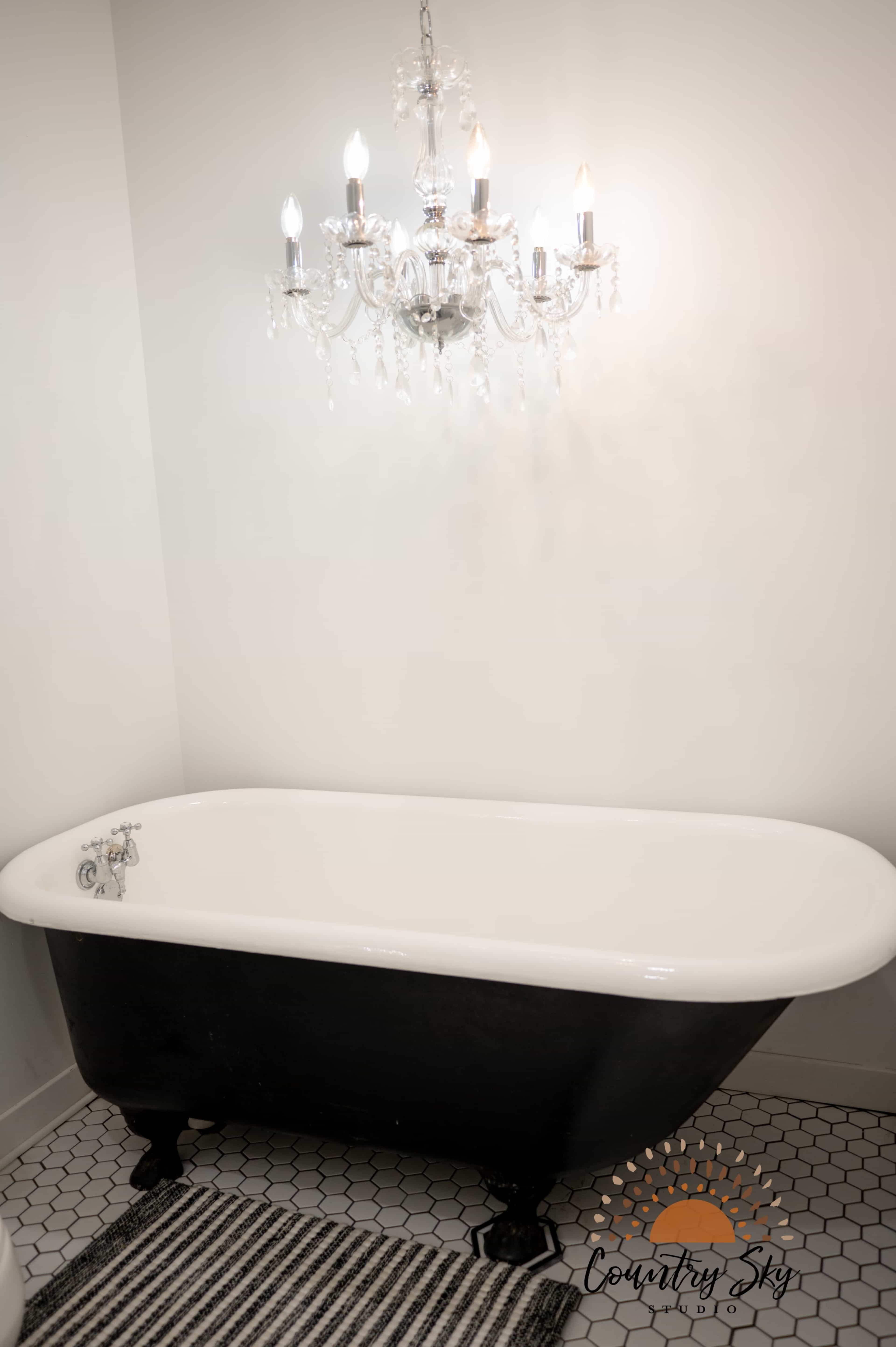 A classic freestanding bathtub with a black exterior and white rim, positioned beneath a crystal chandelier in a bathroom with hexagonal floor tiles.