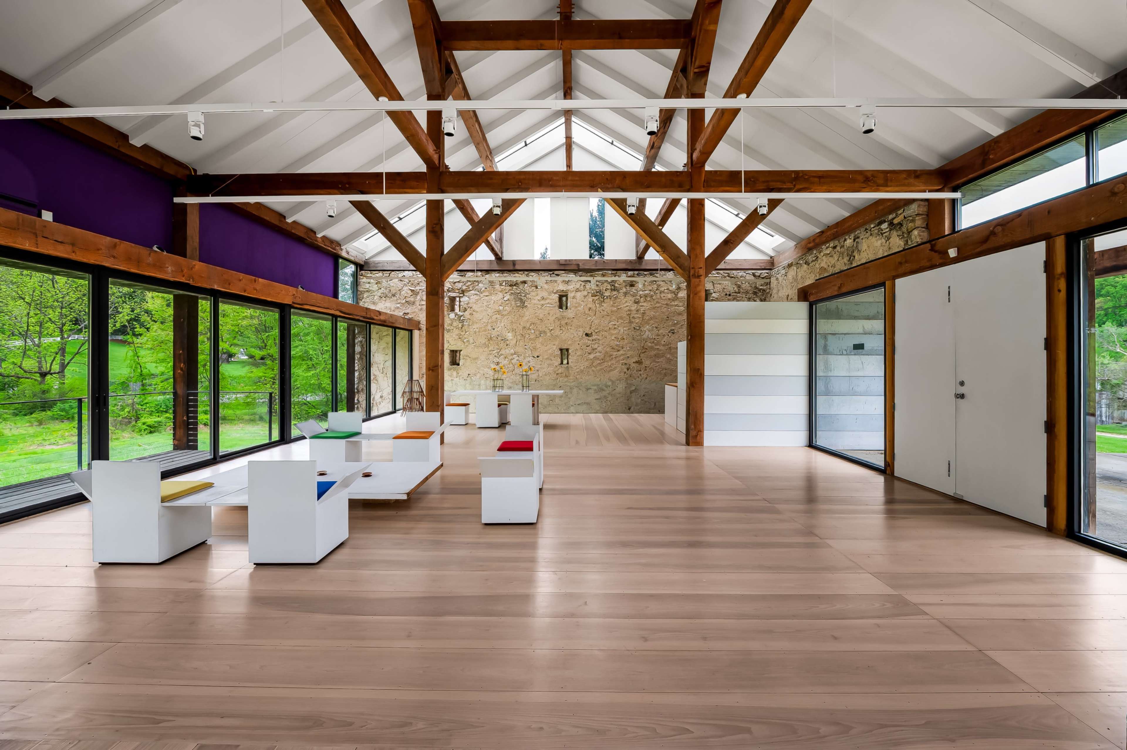 The interior of a modern, spacious room features wooden beams, large windows overlooking greenery, and minimalist furniture in white and colorful accents.