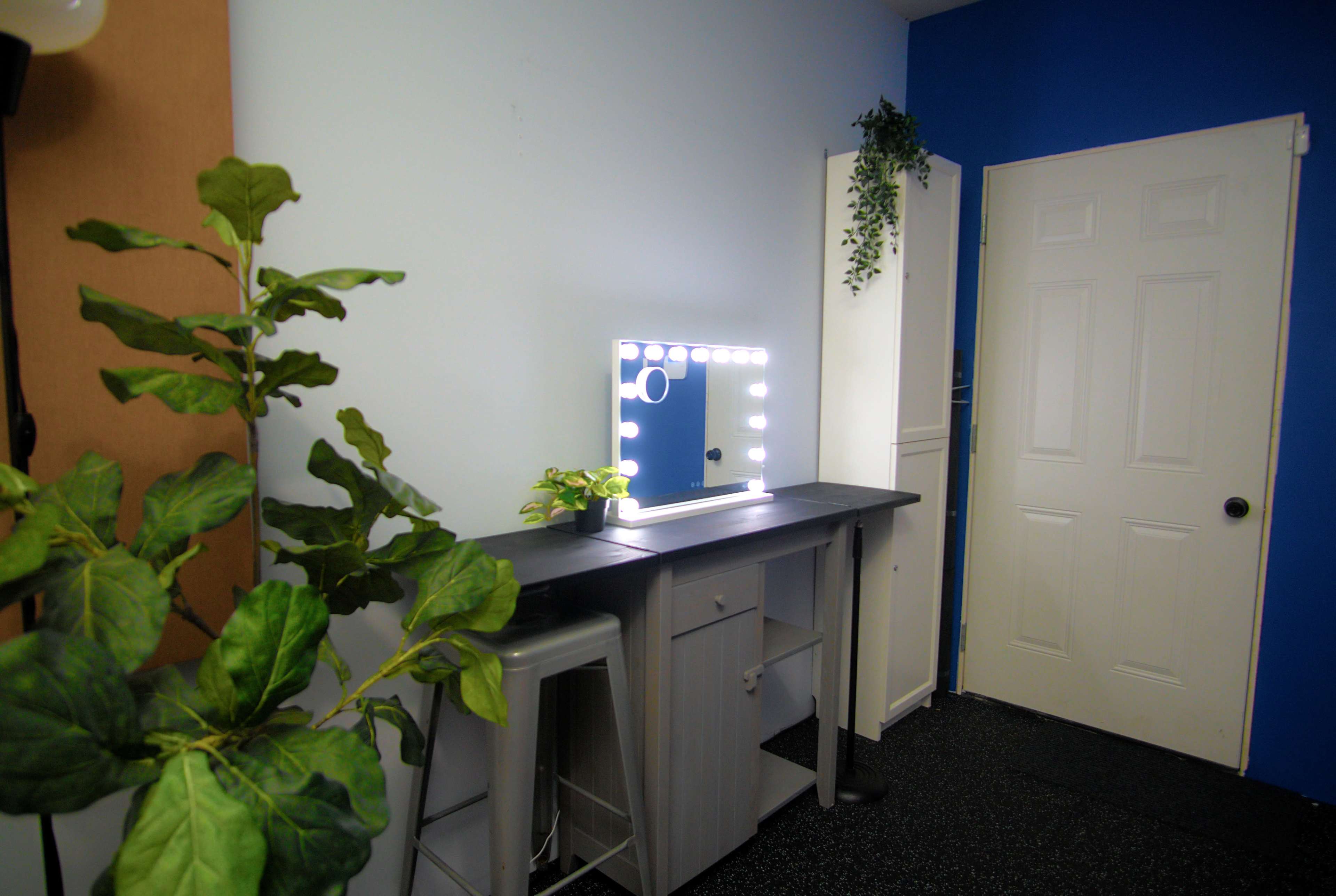 The image shows a well-lit makeup station with a vanity mirror surrounded by light bulbs, a small plant, and a closed white door in a room painted blue and light gray.