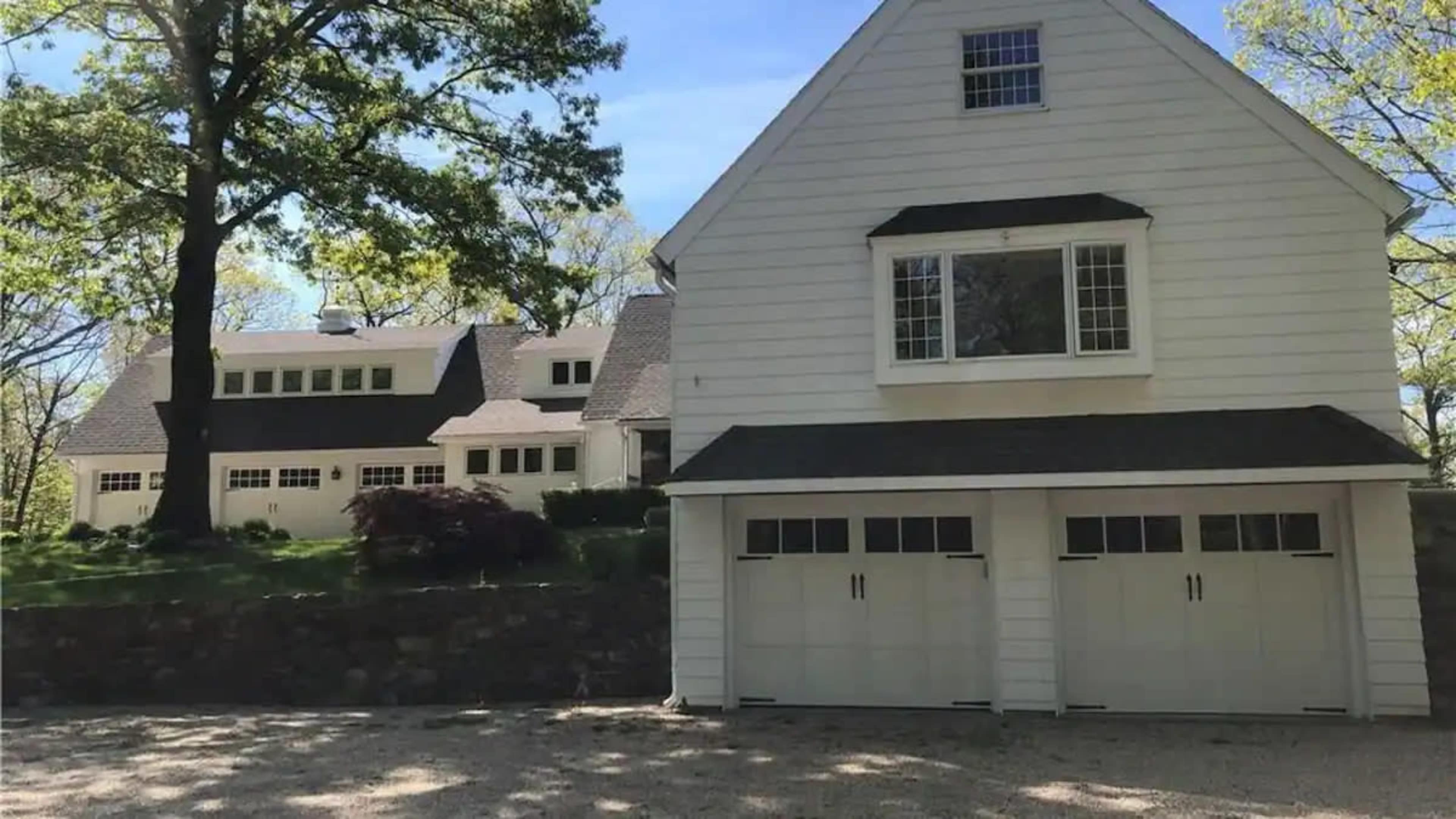 The image shows a two-story white house with a steep roof and attached garage, set in a landscaped area with trees.