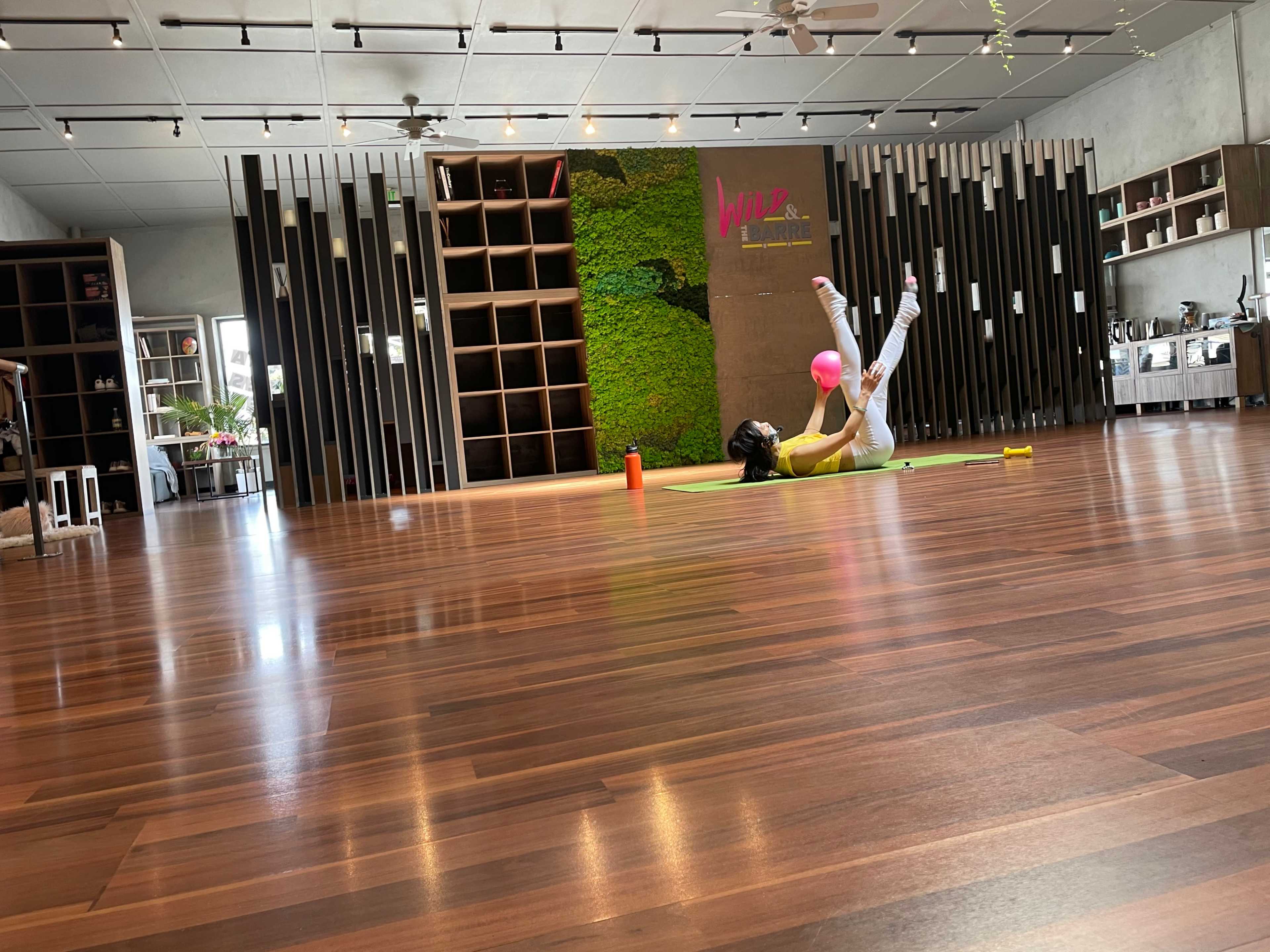 A person is exercising on a green yoga mat in a spacious fitness studio with wooden floors and a green wall feature.