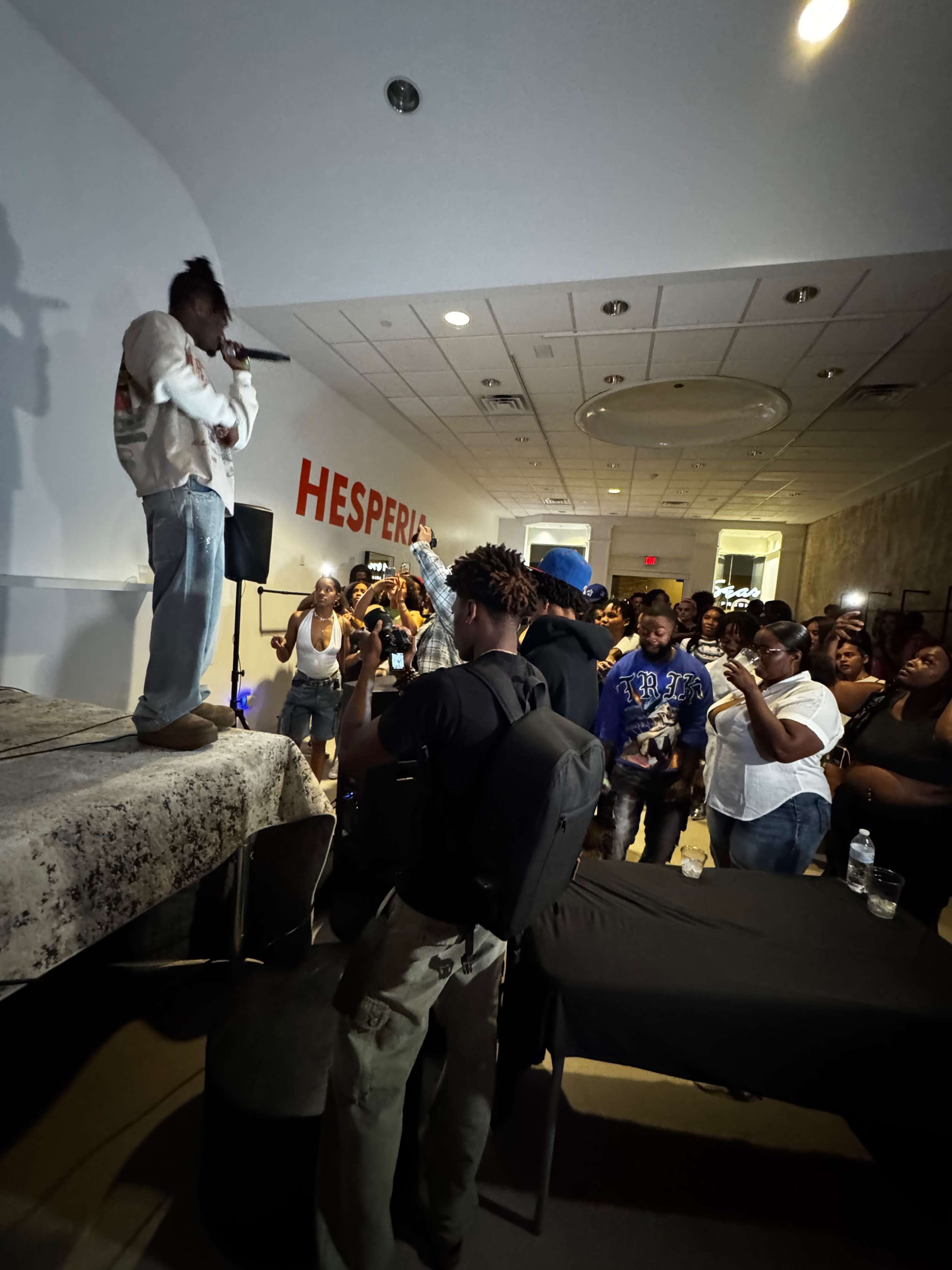 A performer stands on a raised platform while an audience gathers around, capturing the moment with their phones in a dimly lit room.
