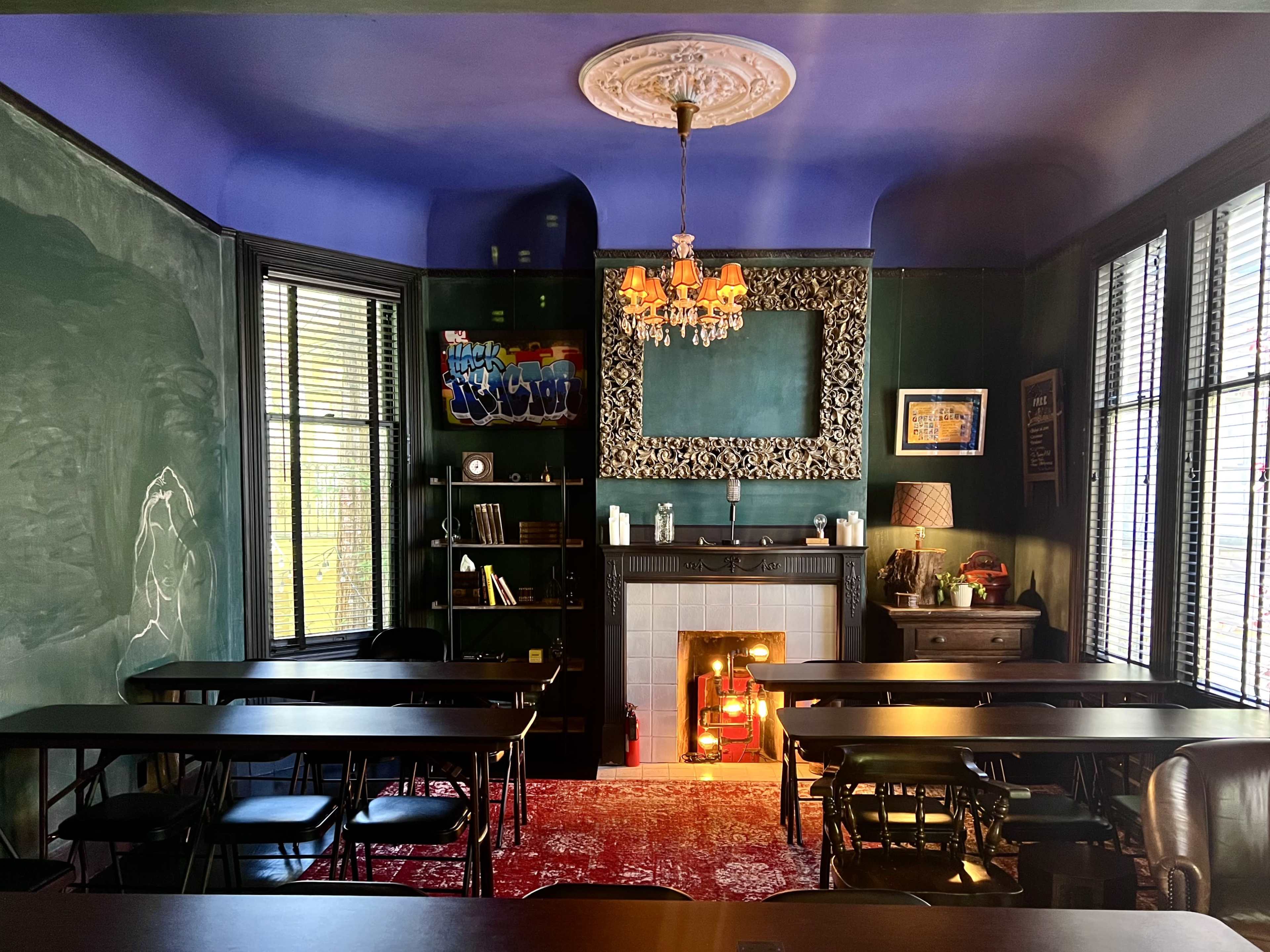 A classroom with dark green walls, a fireplace, and several black tables arranged in rows beneath large windows.