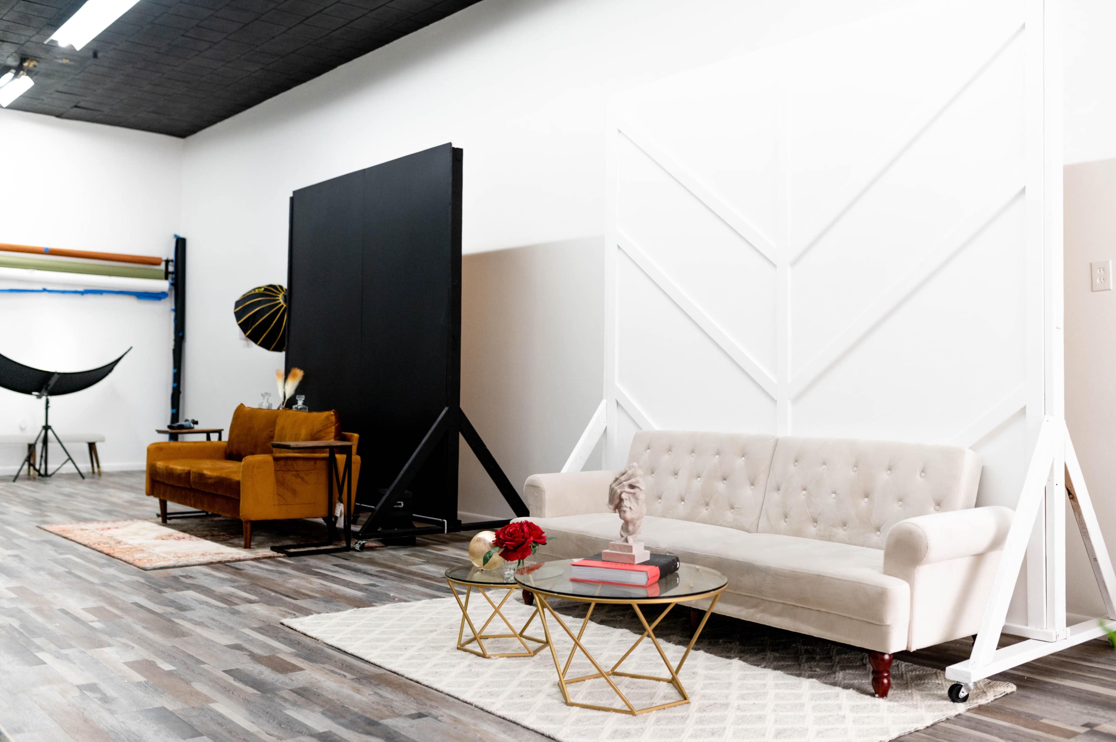 A photography studio featuring a light-colored sofa, a brown couch, a glass coffee table with decorative items, and backdrop setups against a plain wall.