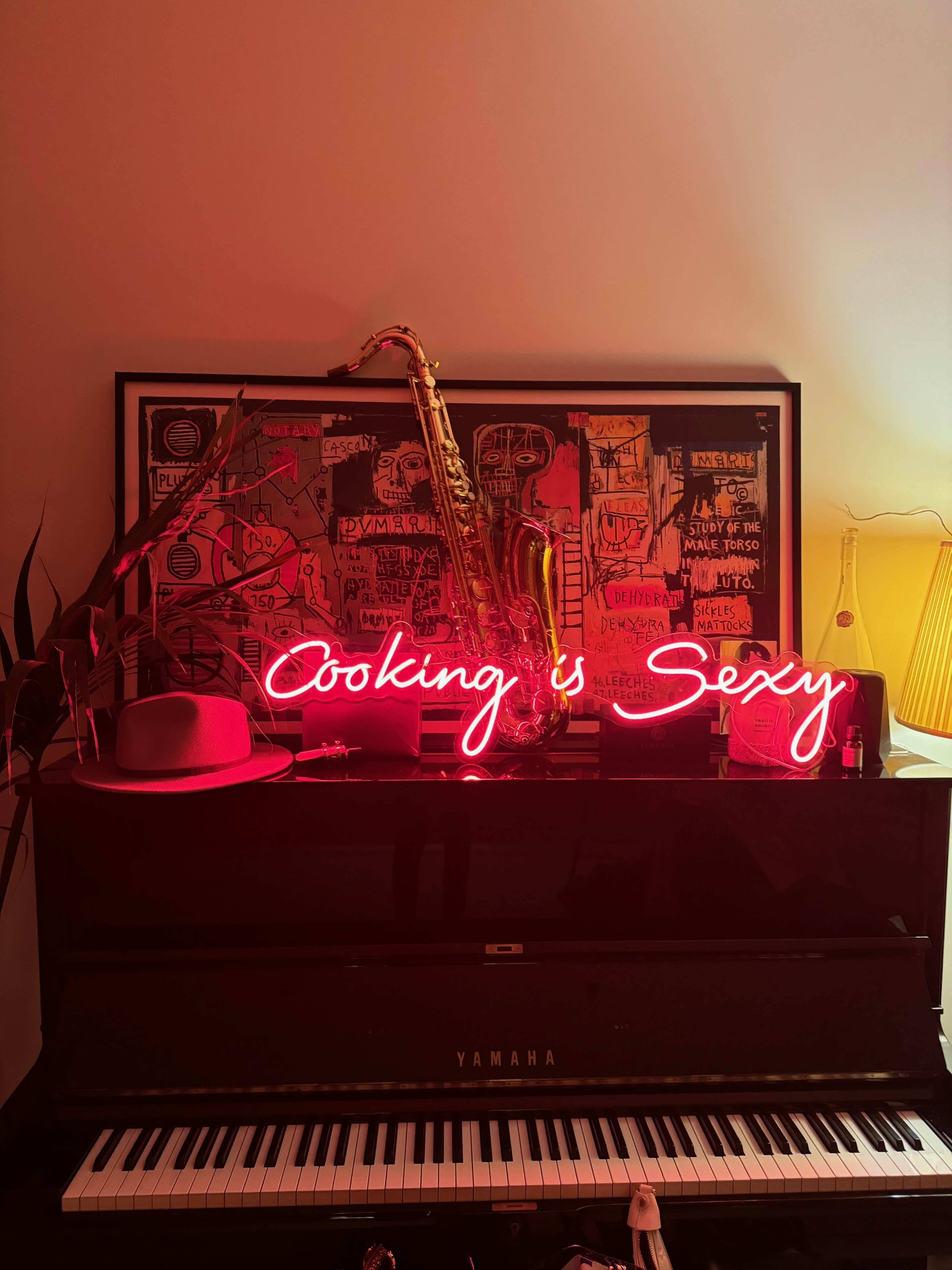 A black piano with a neon sign reading "Cooking is Sexy," a saxophone, and a framed colorful artwork, accompanied by a hat and a lamp.