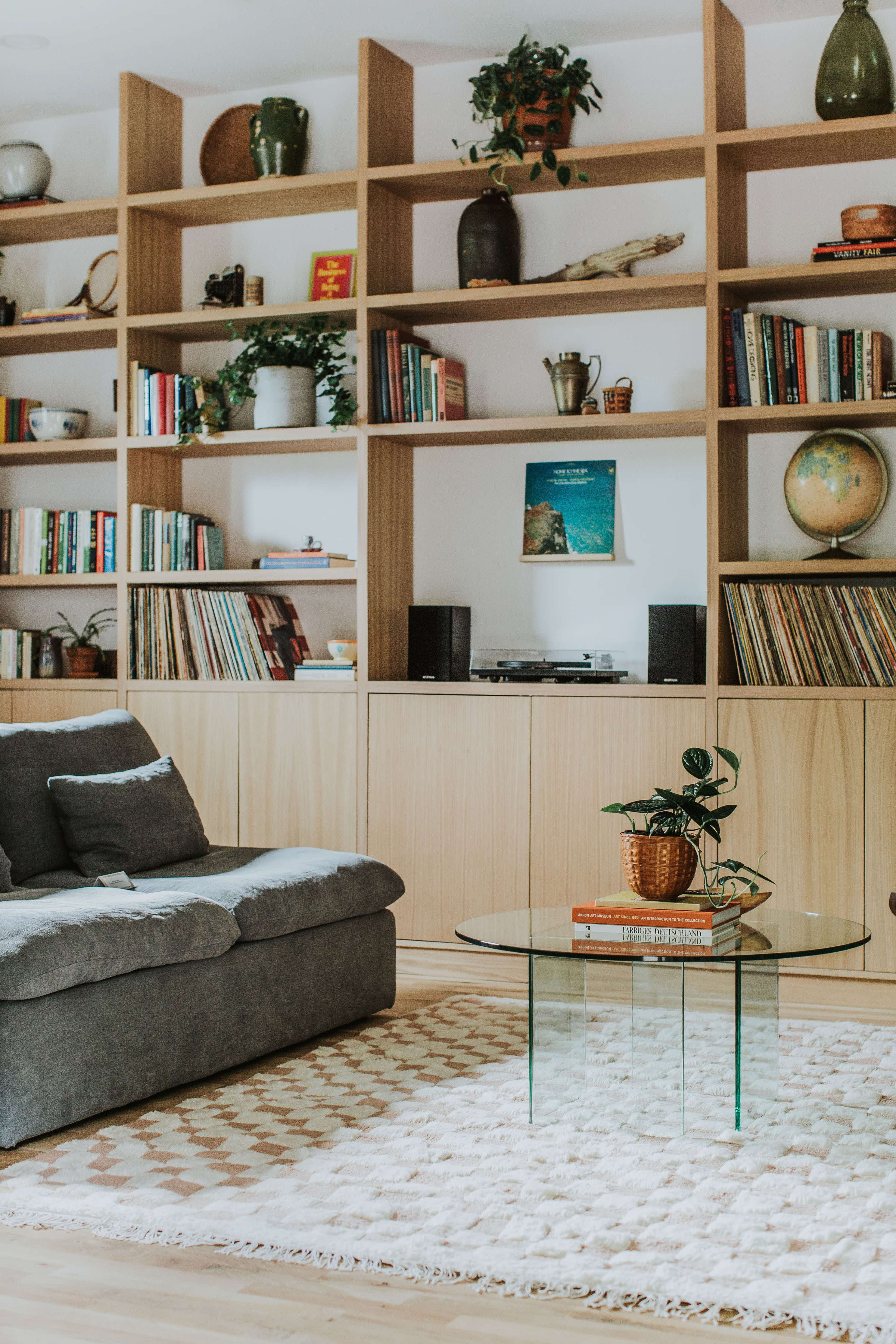 A living room features a gray sofa, a glass coffee table, and shelves filled with books and decorative plants.