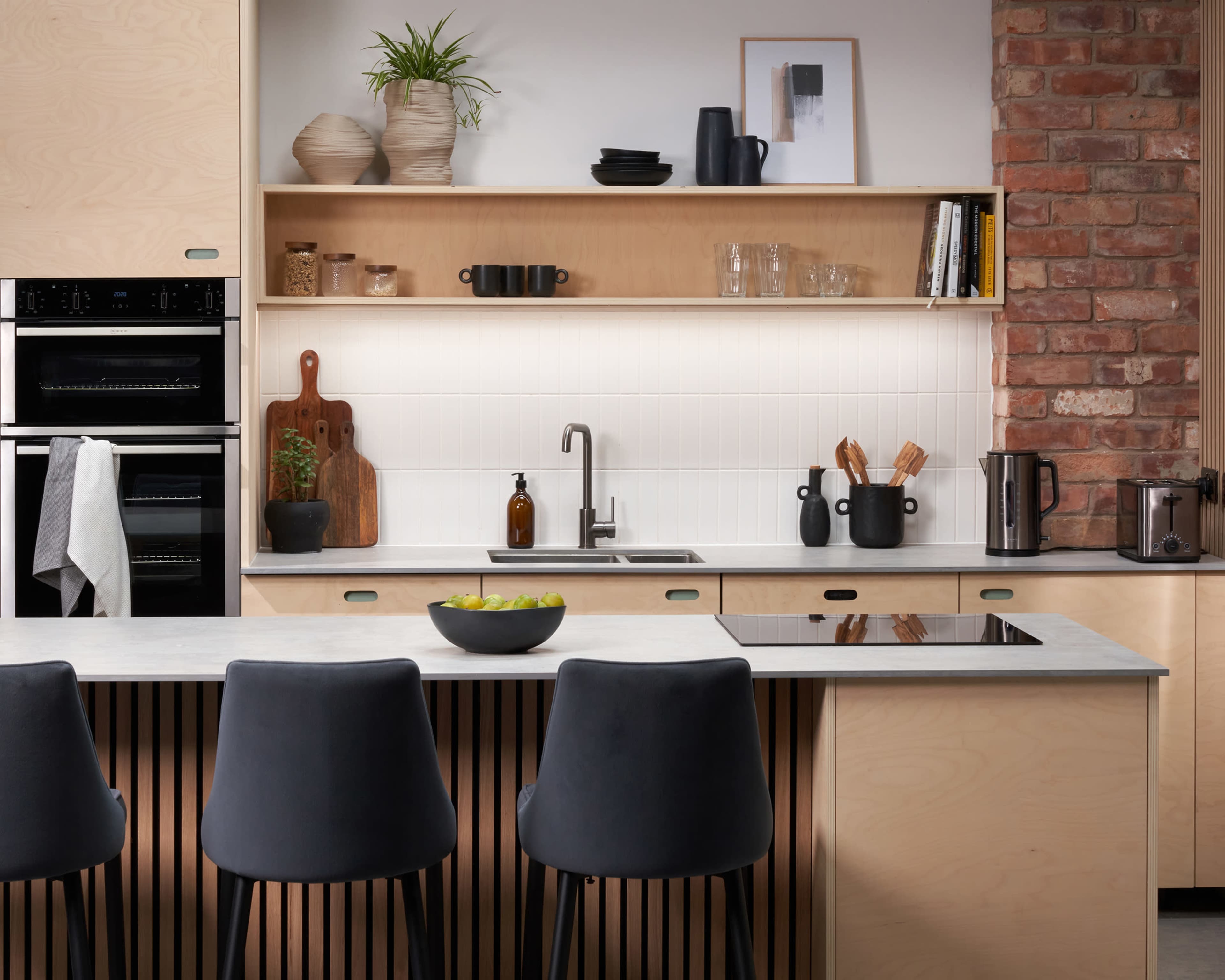 A modern kitchen with a central island, featuring wooden cabinetry, black stools, and various kitchen utensils arranged on the countertops.