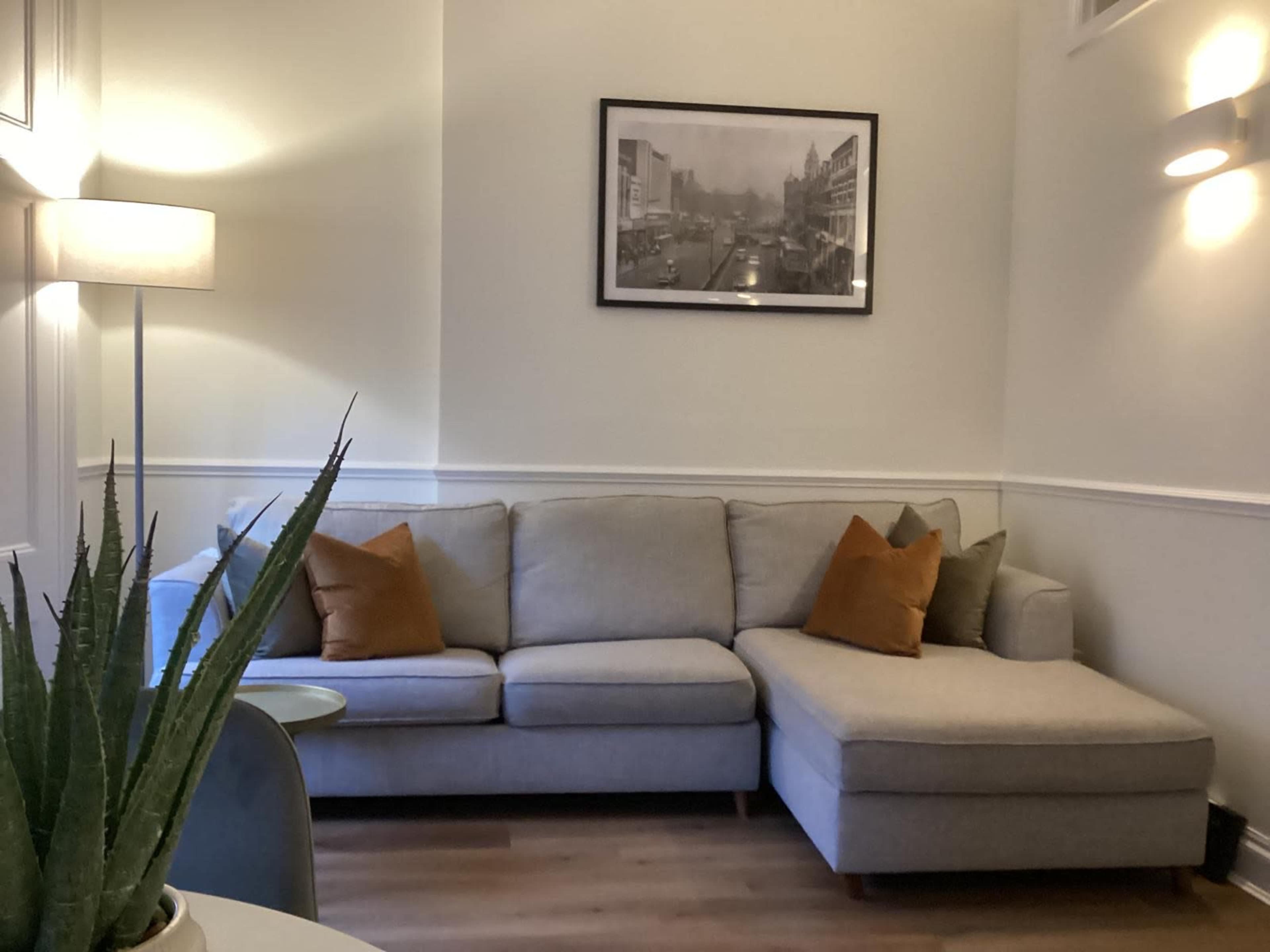 The image shows a cozy living room corner featuring a gray sectional couch with orange accent pillows, a small round table, an indoor plant, and a framed black-and-white photograph on the wall.