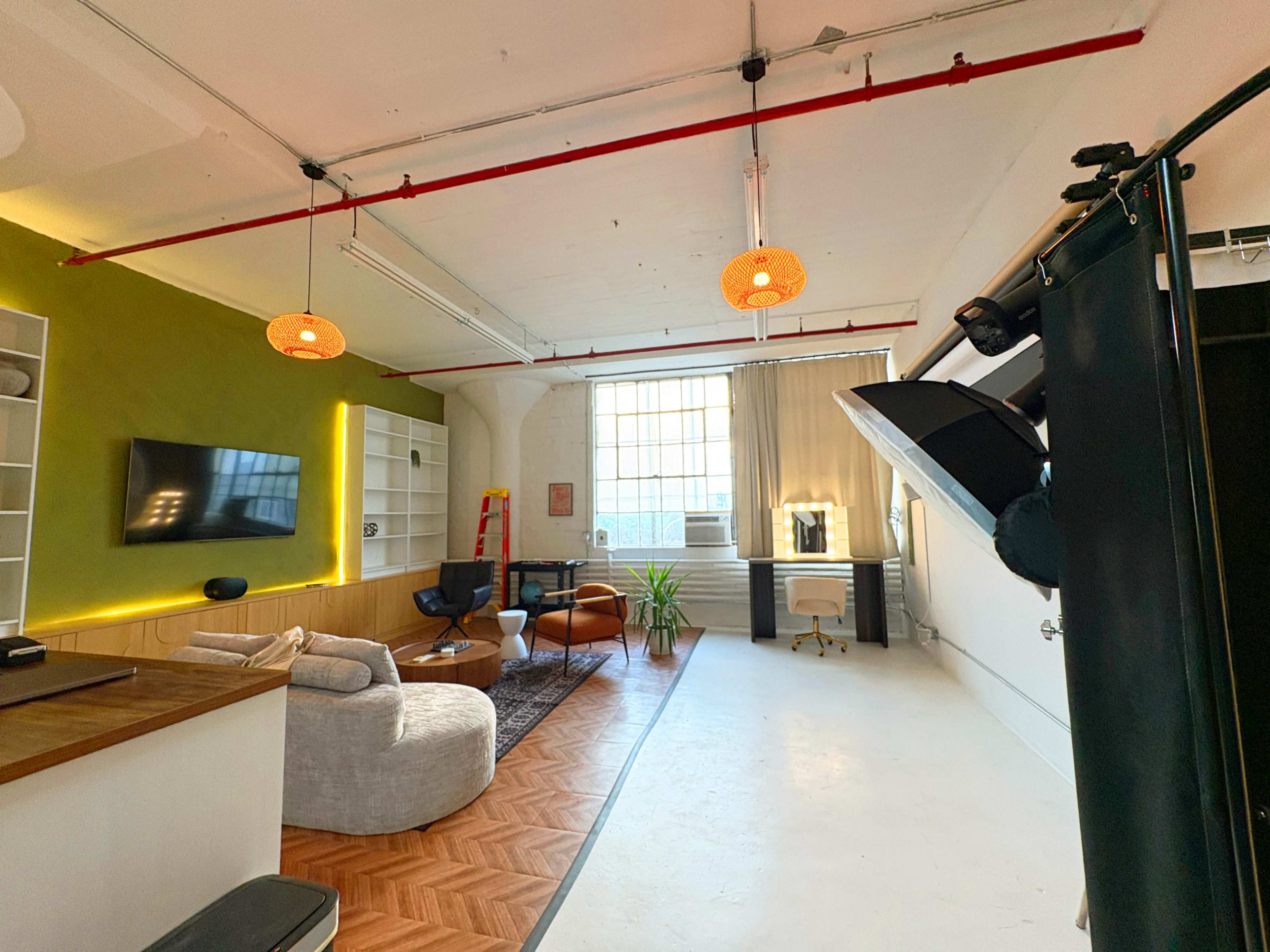 The image shows a well-lit studio apartment with a green accent wall, a seating area, a large window, and minimalistic decor.