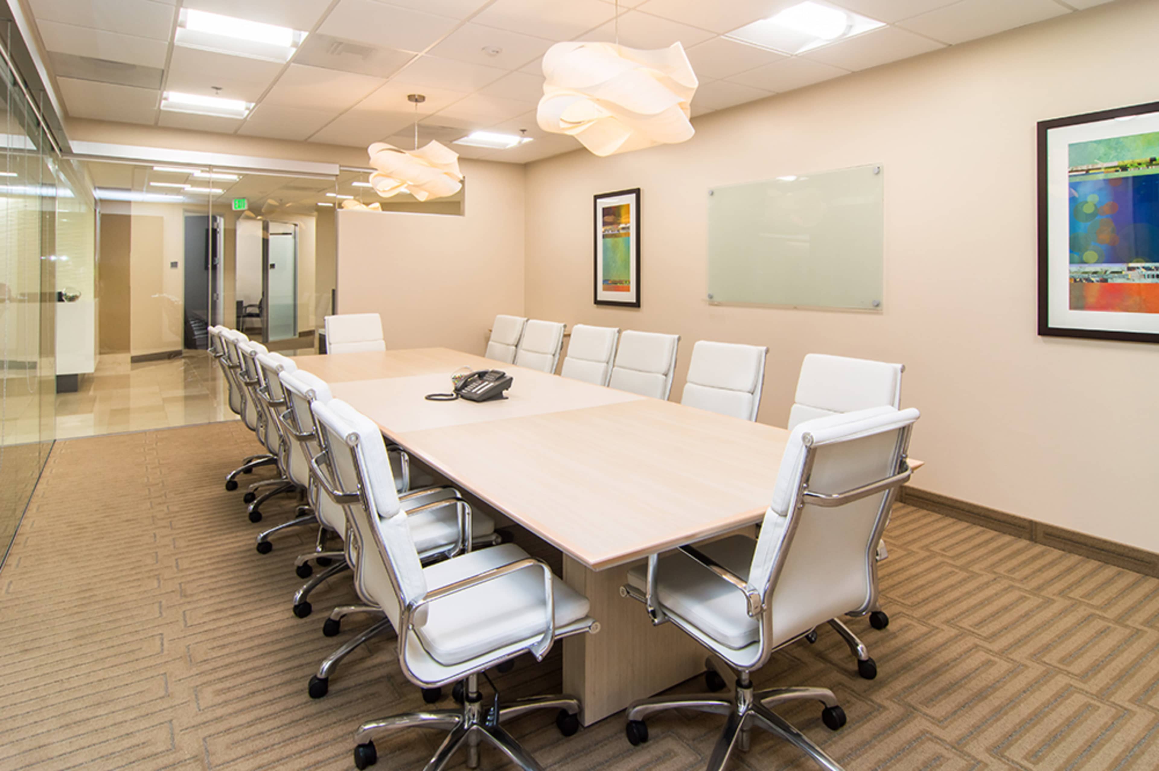 A long conference table with white chairs is set up in a well-lit meeting room featuring modern lighting and artwork on the walls.