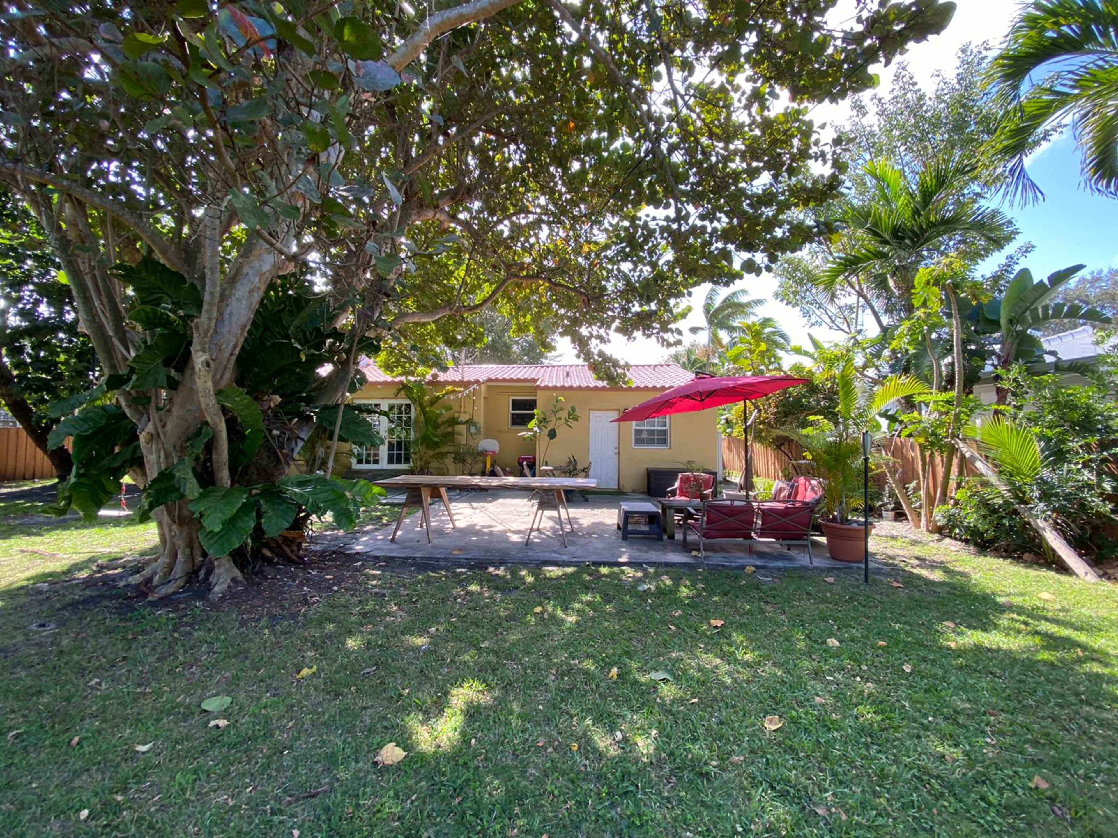 A yellow house with a red umbrella is surrounded by lush greenery and a wooden deck in a backyard.